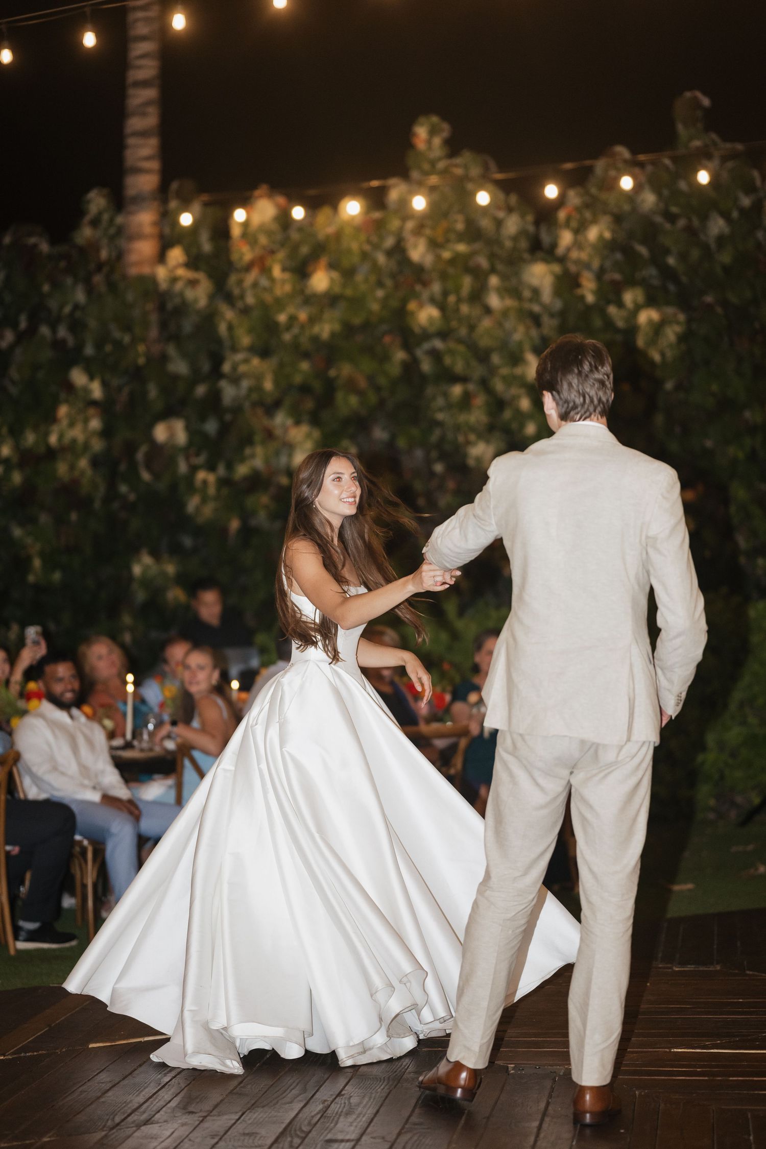 Wedding couple dancing under string lights on wooden deck at night.