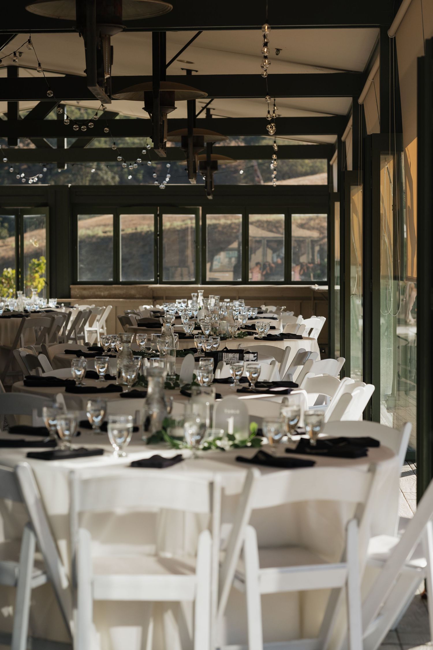 Elegant wedding reception setup with white tablecloths and chairs in modern venue with glass walls.