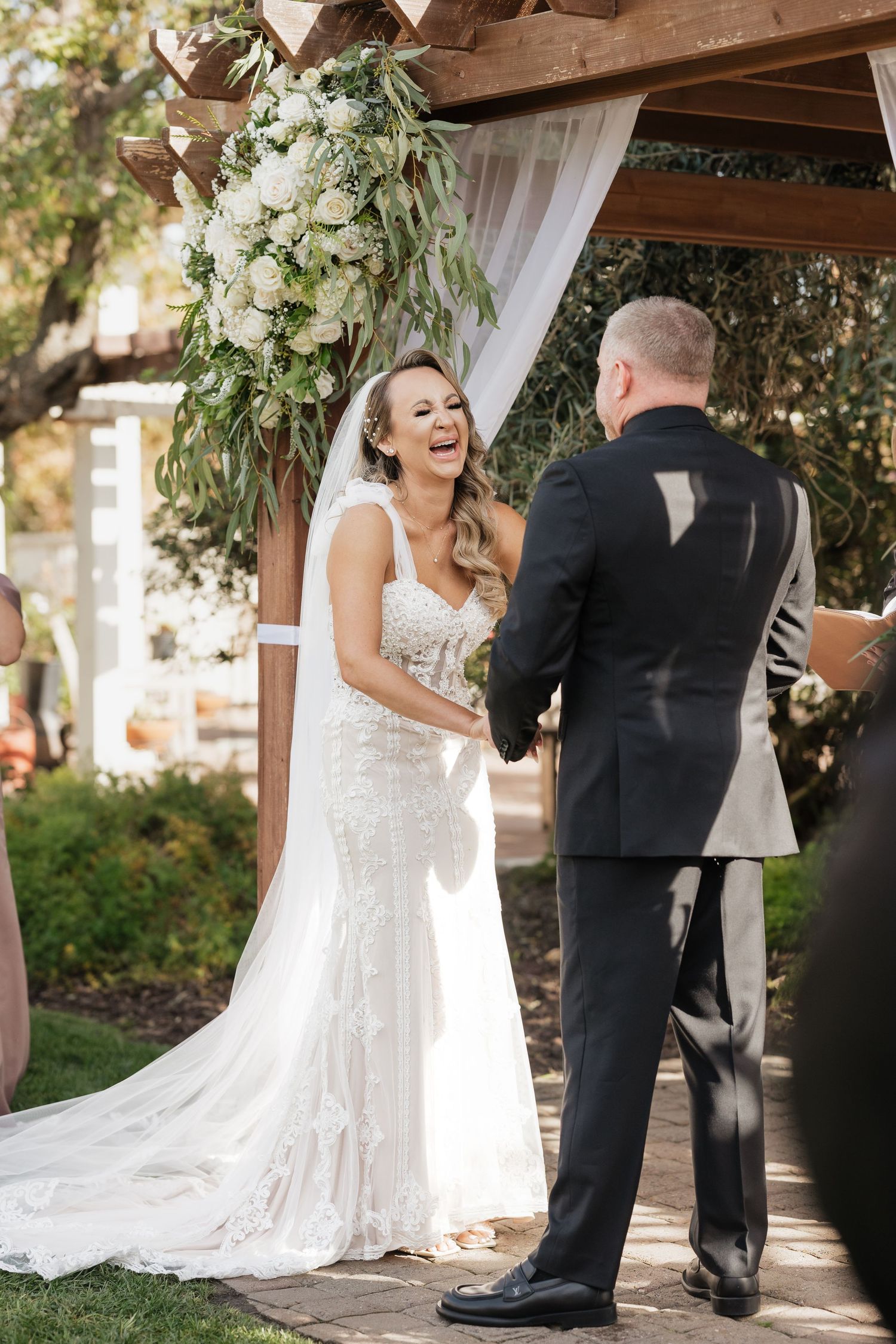 Wedding ceremony moment under wooden arch decorated with greenery and white fabric.