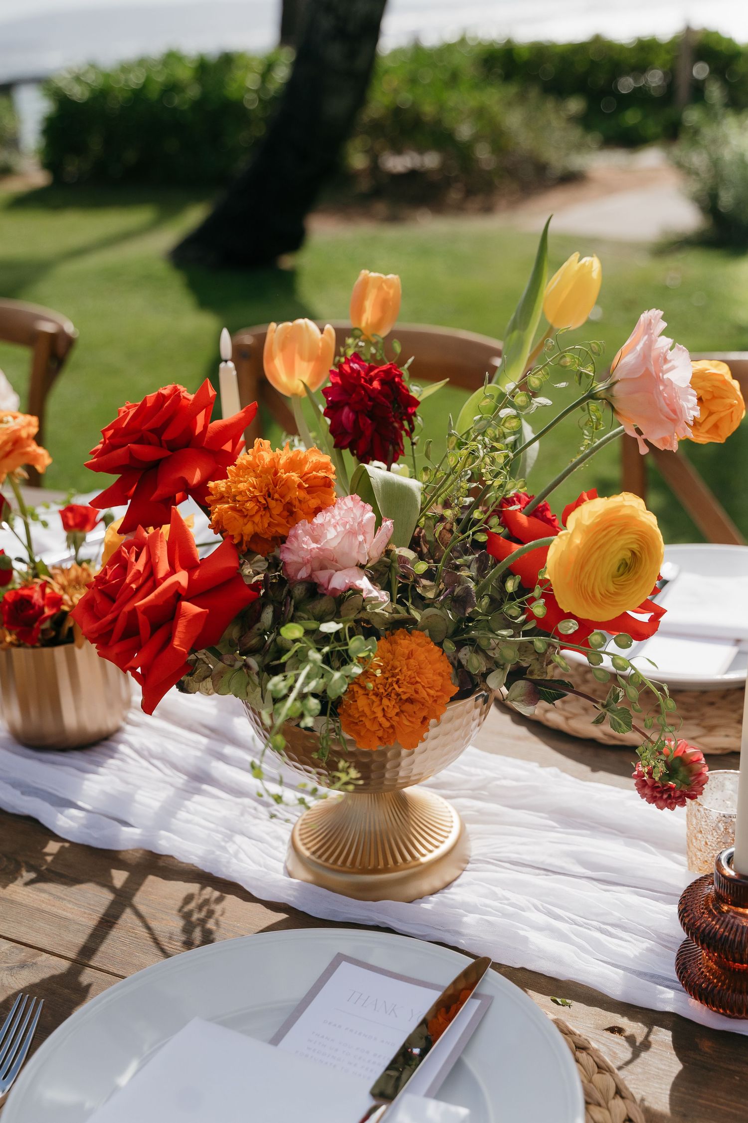 Colorful floral centerpieces with red, orange and pink flowers in gold vases decorate a white tablecloth outdoors.