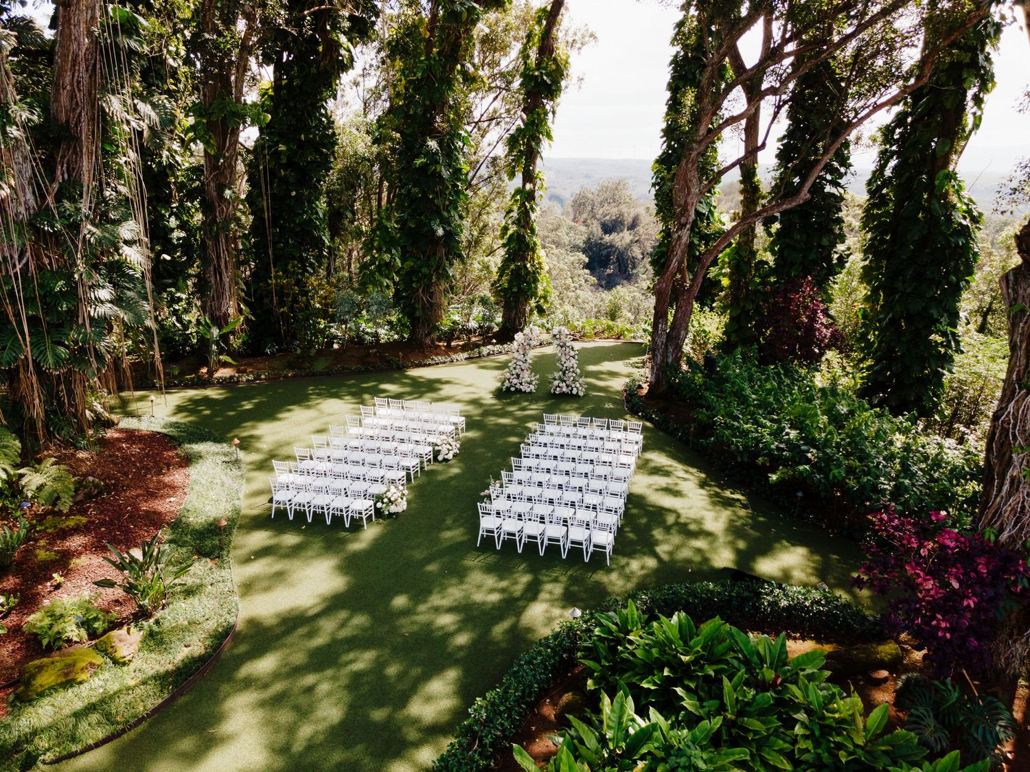 Outdoor wedding ceremony setup with white chairs on green lawn surrounded by tall trees and lush tropical gardens.