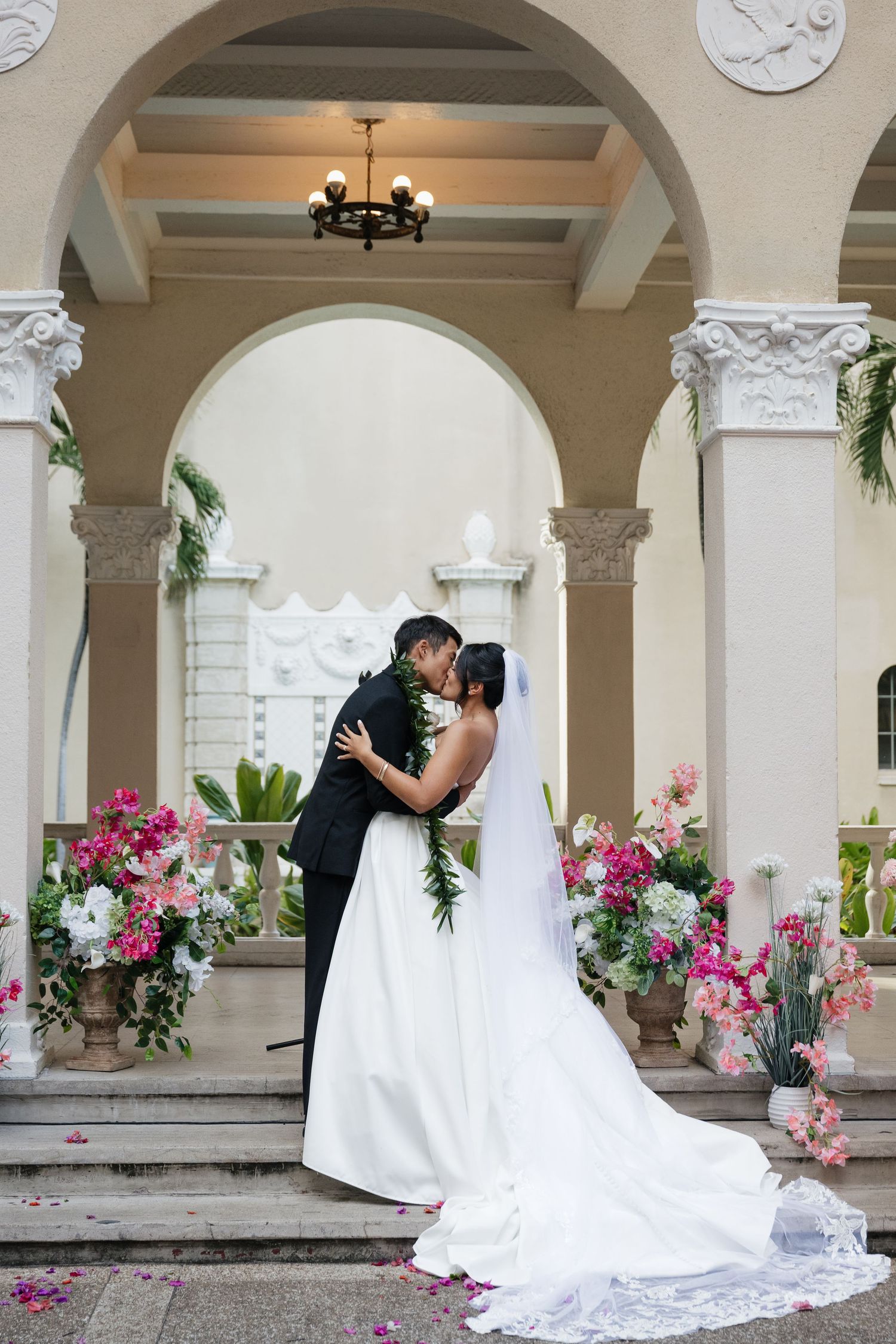 Bride and groom share first kiss at elegant venue with pink floral arrangements and grand arched columns.