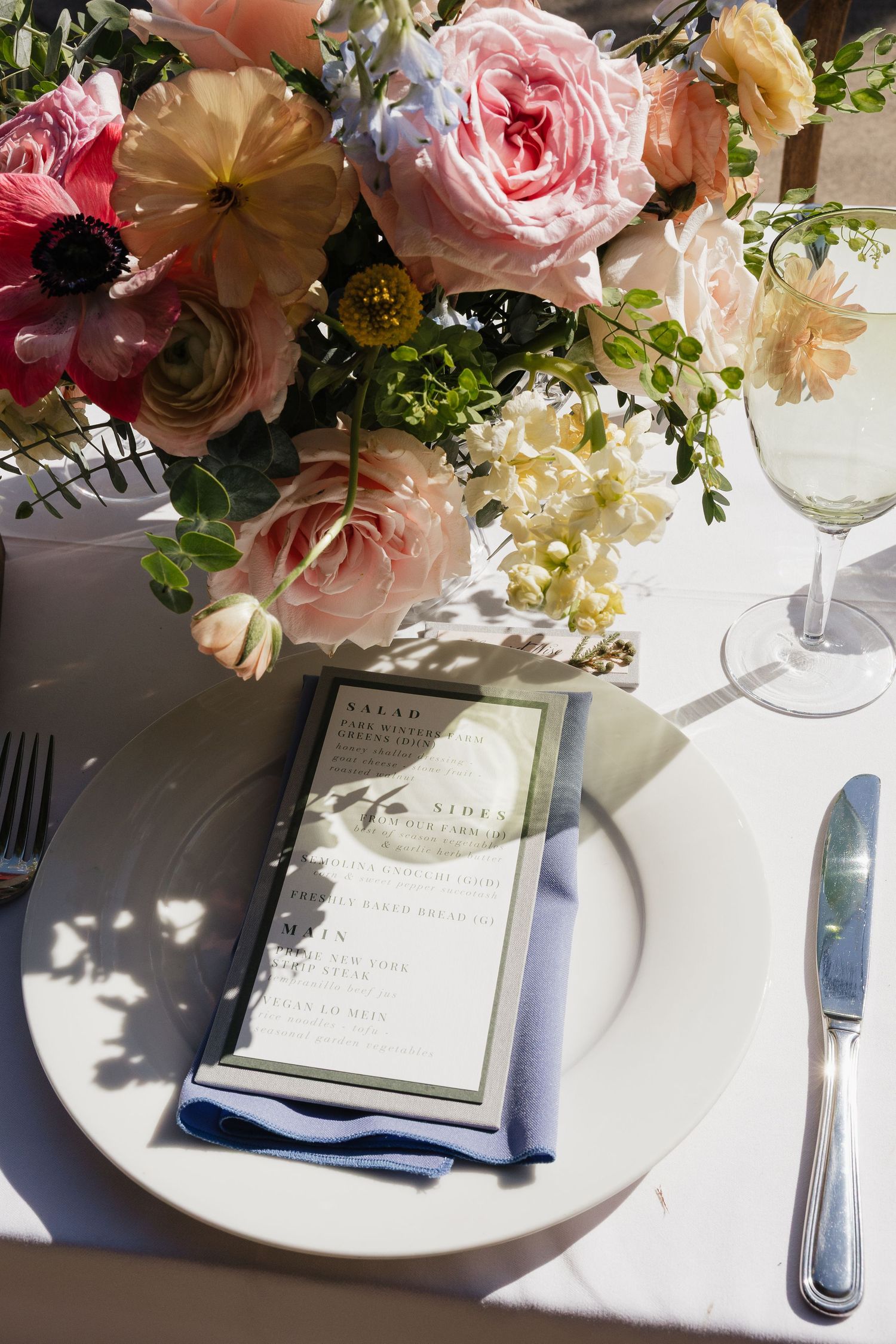 Elegant table setting with pink and peach floral centerpiece, white plates, and menu cards in natural sunlight.