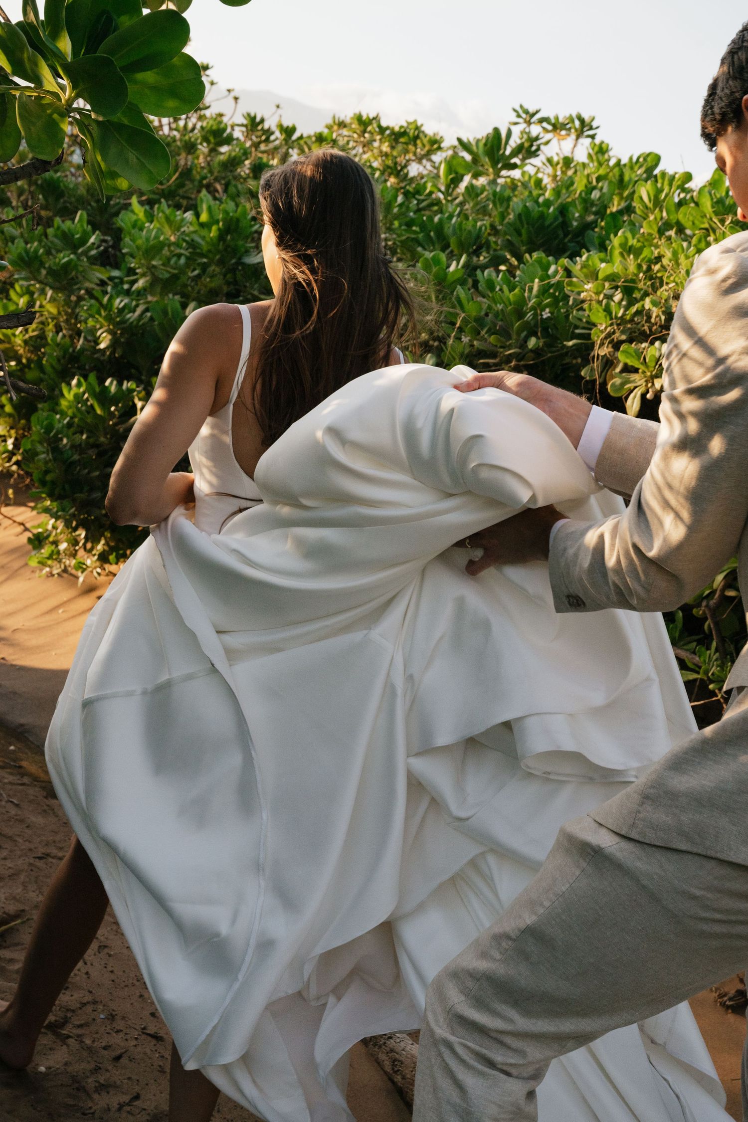 A flowing white wedding dress billows in the wind against lush tropical greenery at sunset.
