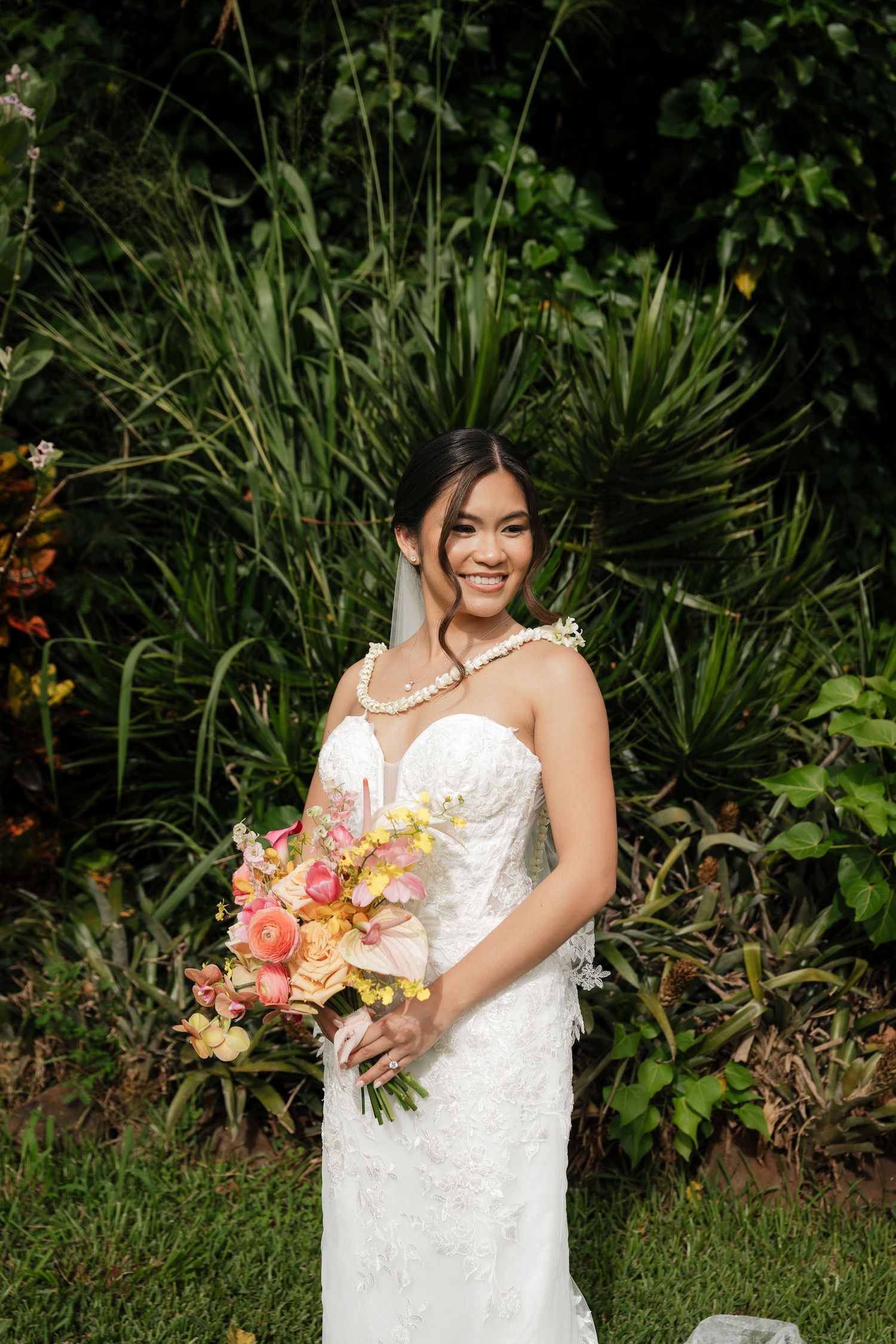 Smiling bride in strapless lace gown holding colorful tropical bouquet in lush garden setting.