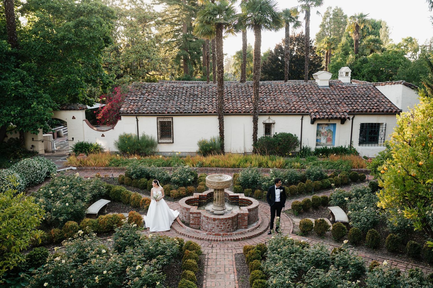 Bride and groom stand in a lush garden courtyard with fountain beside a white Spanish-style villa.
