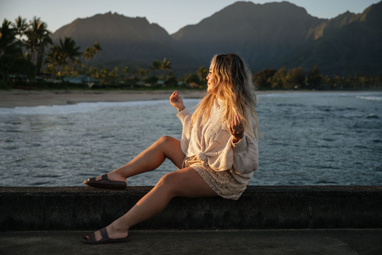 Person in white sweater sits by ocean at sunset with mountains and palm trees in background in Hanalei Bay, Hawaii.