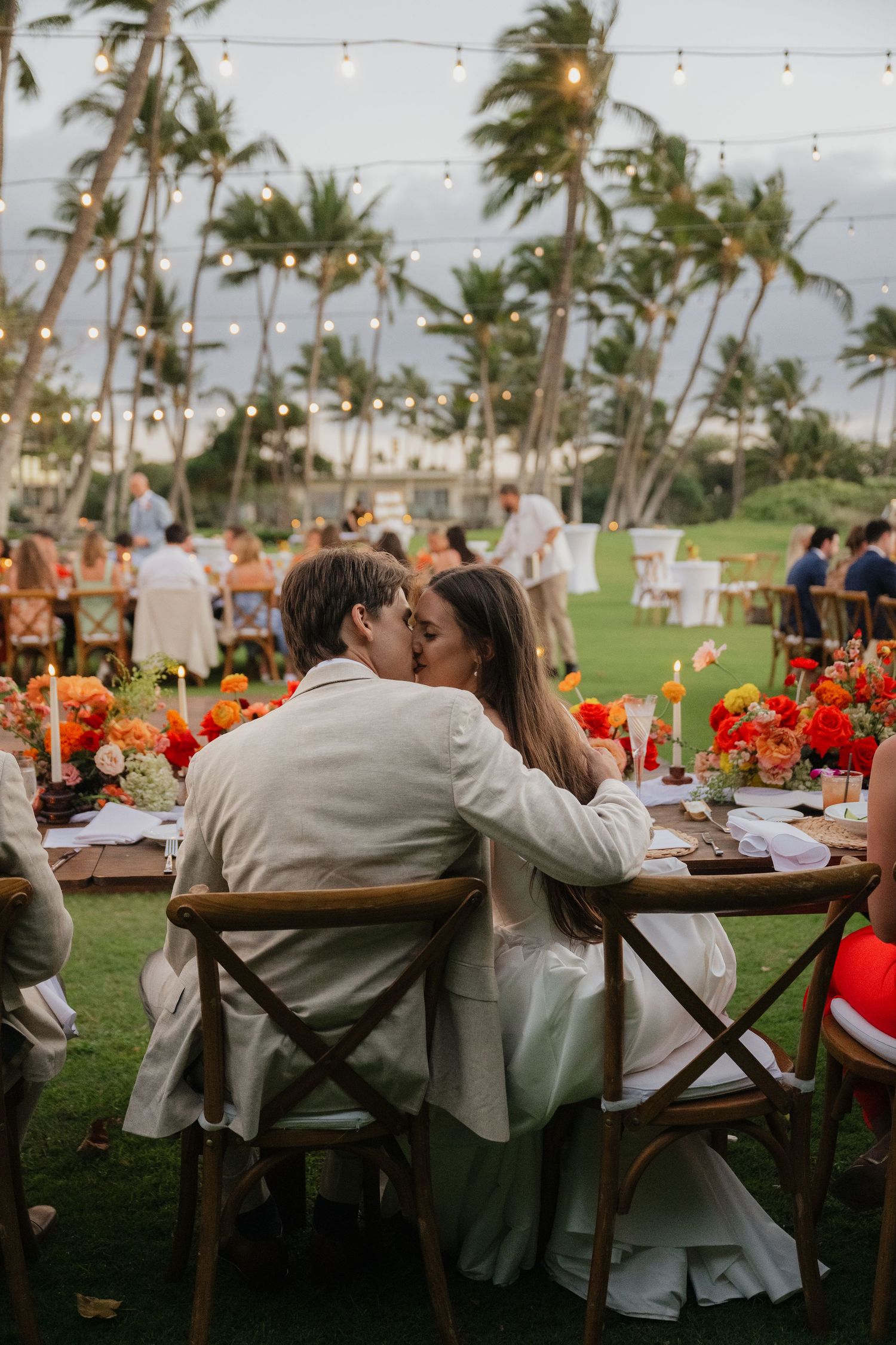 Outdoor wedding reception under string lights with palm trees and orange floral decorations on rustic wooden tables.