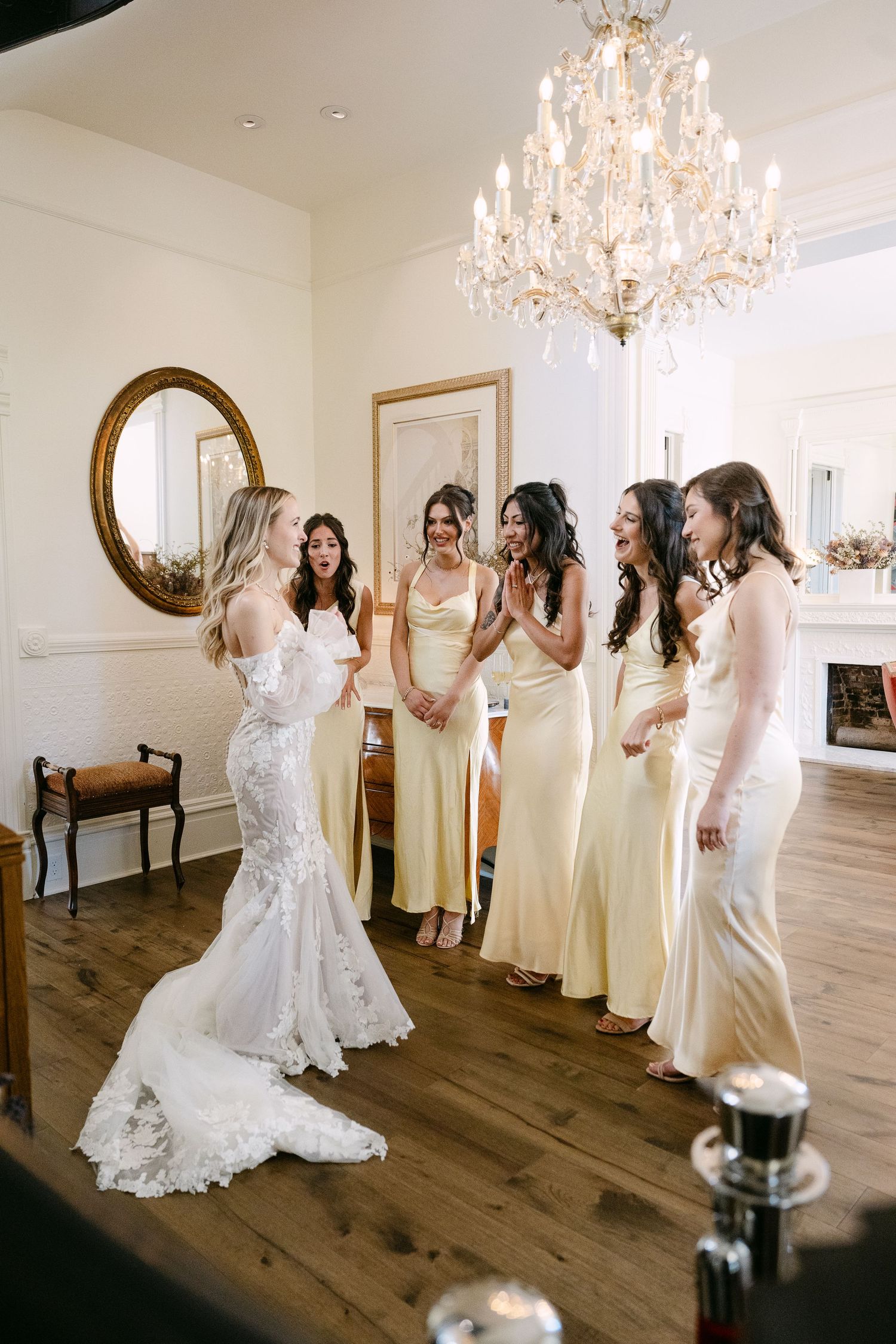 Bridesmaids in cream colored dresses gather around a bride in white lace for candid getting ready photos.