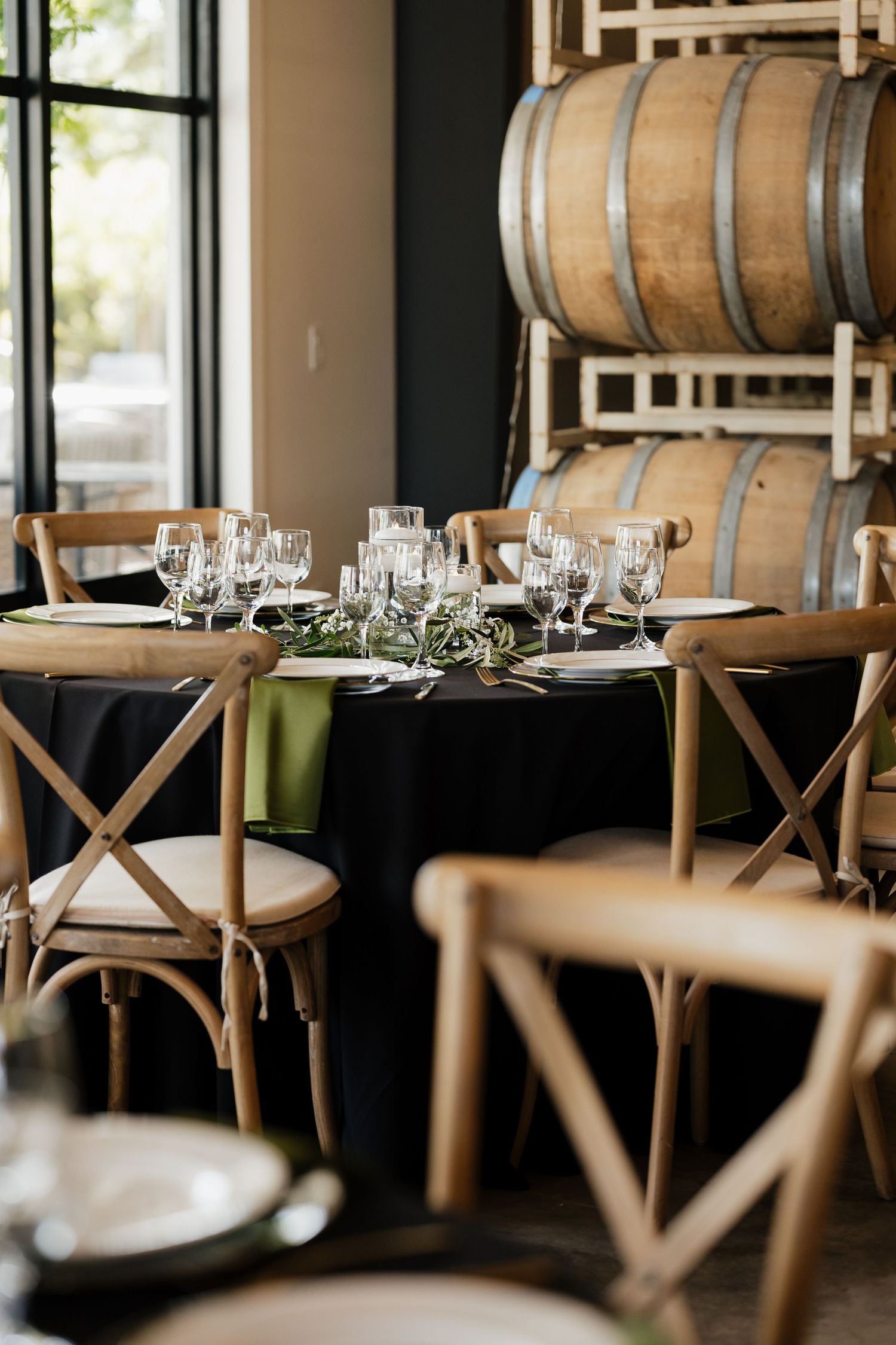 Elegant winery event setup with wooden cross-back chairs, black tablecloths, wine glasses, and oak barrels in the background.