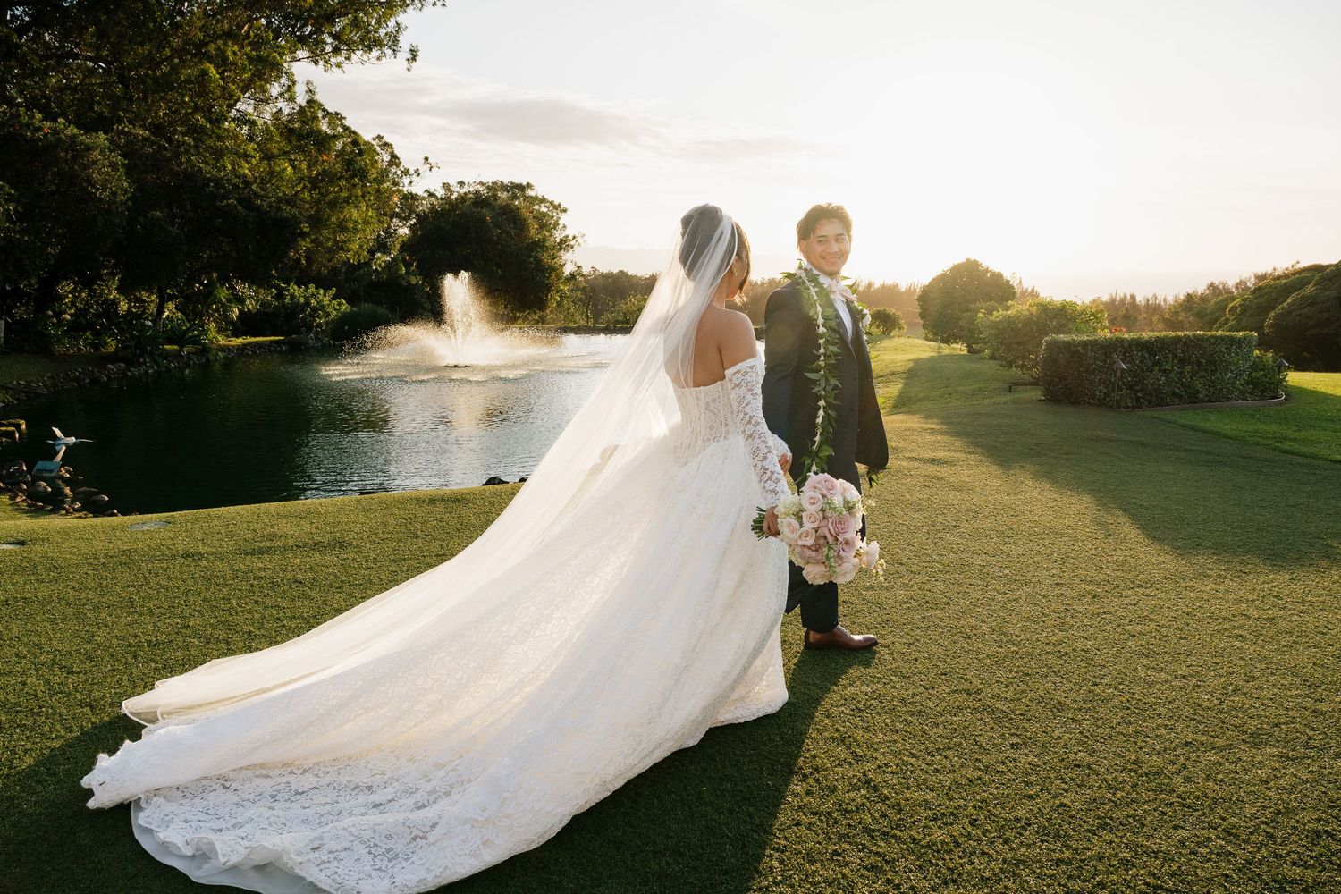 Bride and groom walking hand in hand on lush green lawn at sunset with fountain and pond in background.