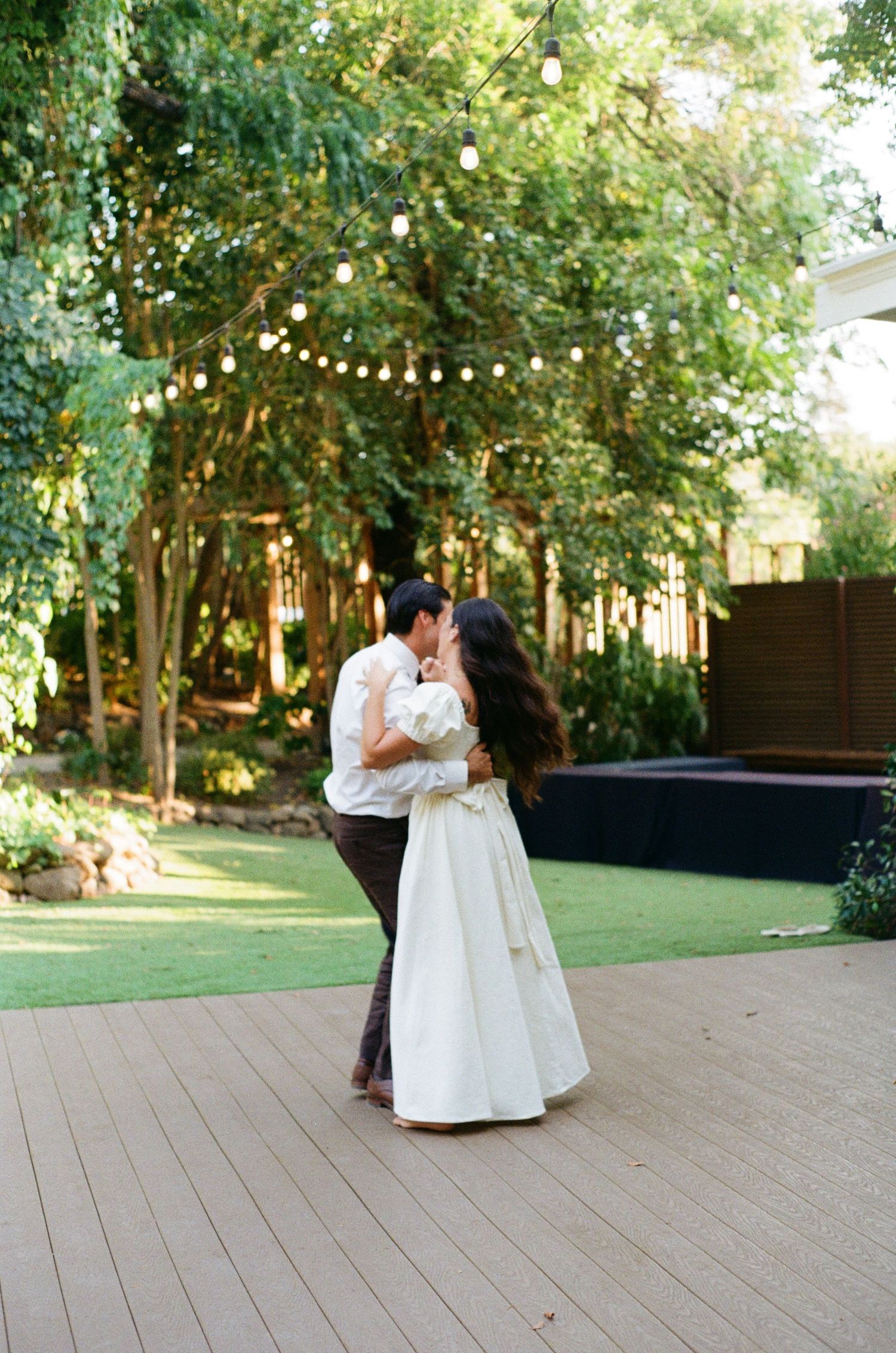 A romantic moment captured on an outdoor deck with string lights and trees creating an intimate garden setting.