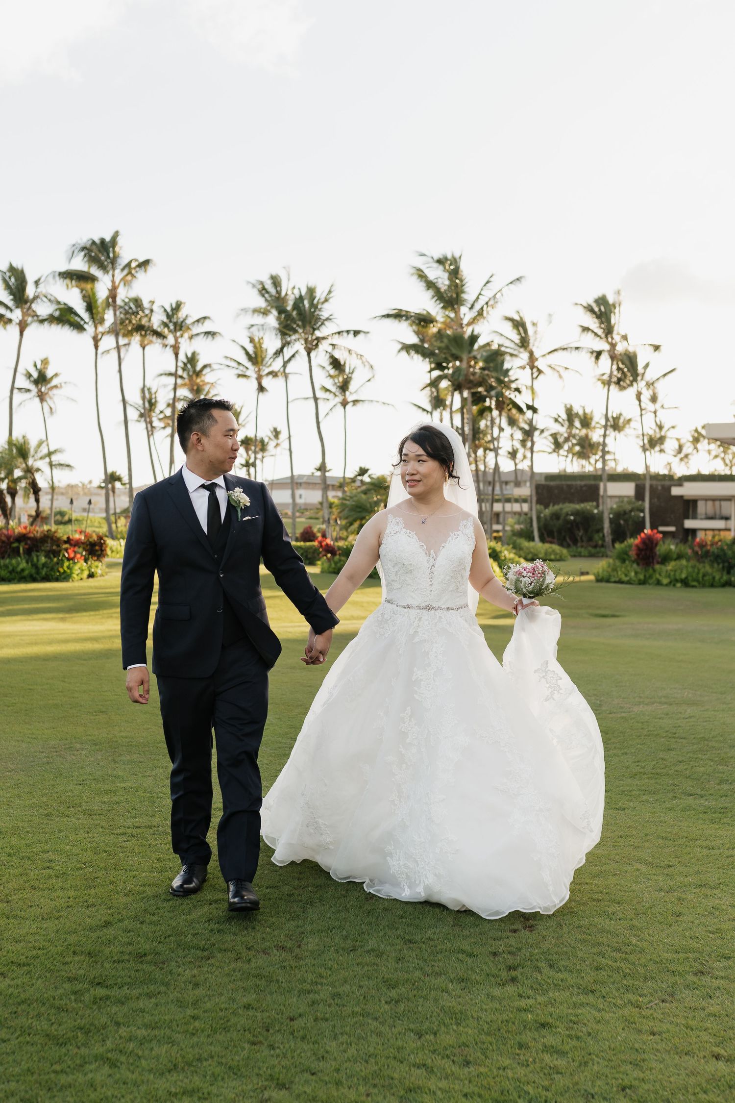 Wedding couple walks hand in hand across a manicured lawn with tropical palm trees.