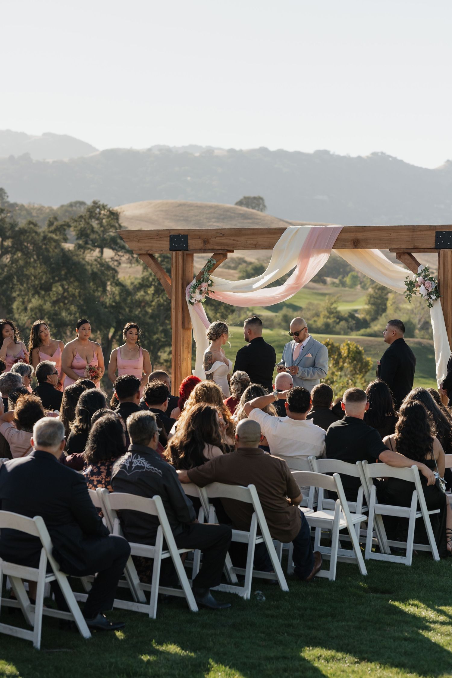 Outdoor wedding ceremony with white chairs and wooden arch overlooking mountain landscape.