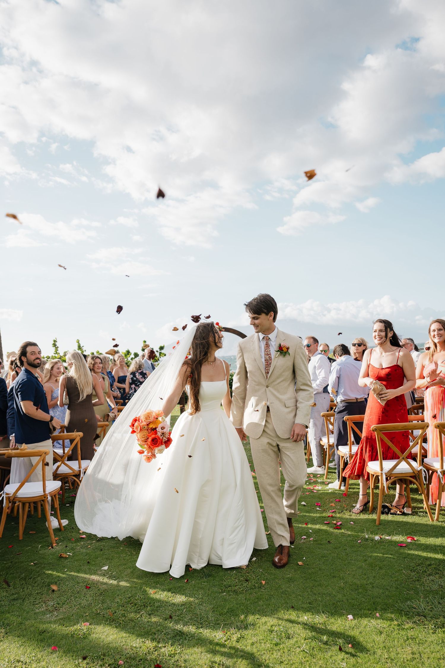 Wedding guests throw flower petals as newlyweds celebrate on a scenic green lawn.