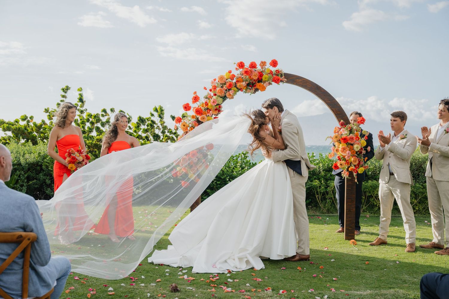 A bride in a flowing white dress poses with red balloons in a scenic outdoor garden setting while her veil catches the breeze.