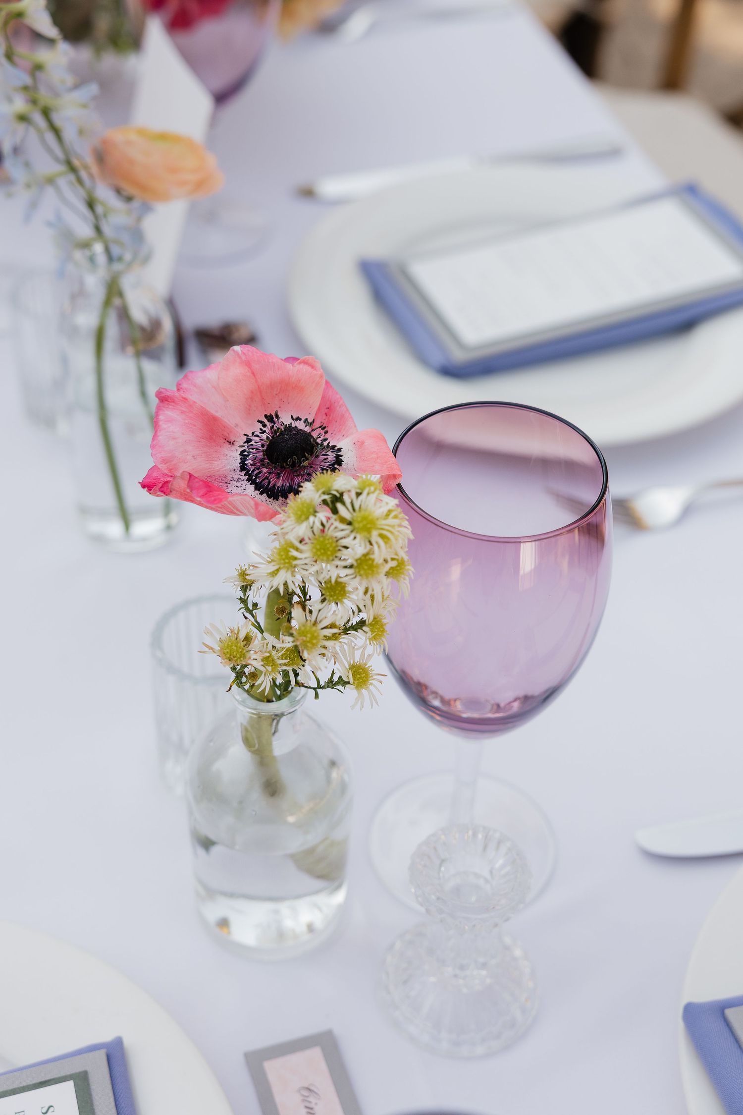 Elegant table setting with pink anemone flowers and daisies in small vases next to purple glassware on white tablecloth.