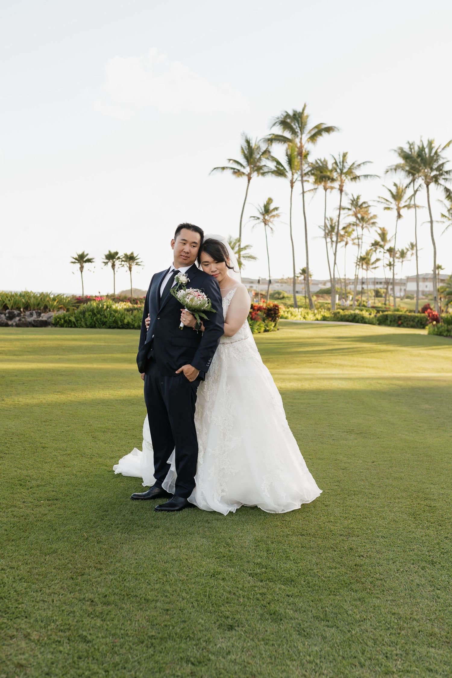 Wedding couple poses together on a lush green lawn with palm trees in the background.