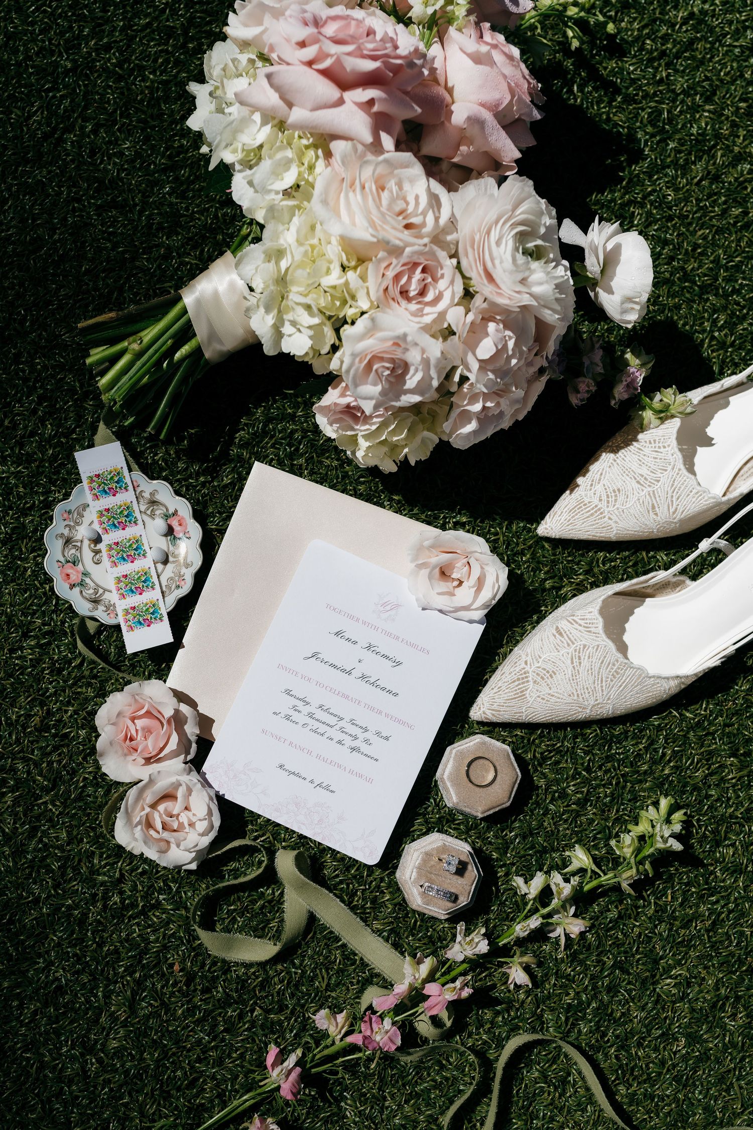 Elegant wedding flat lay with blush pink bouquet, lace heels, invitation, and floral accessories on green grass.