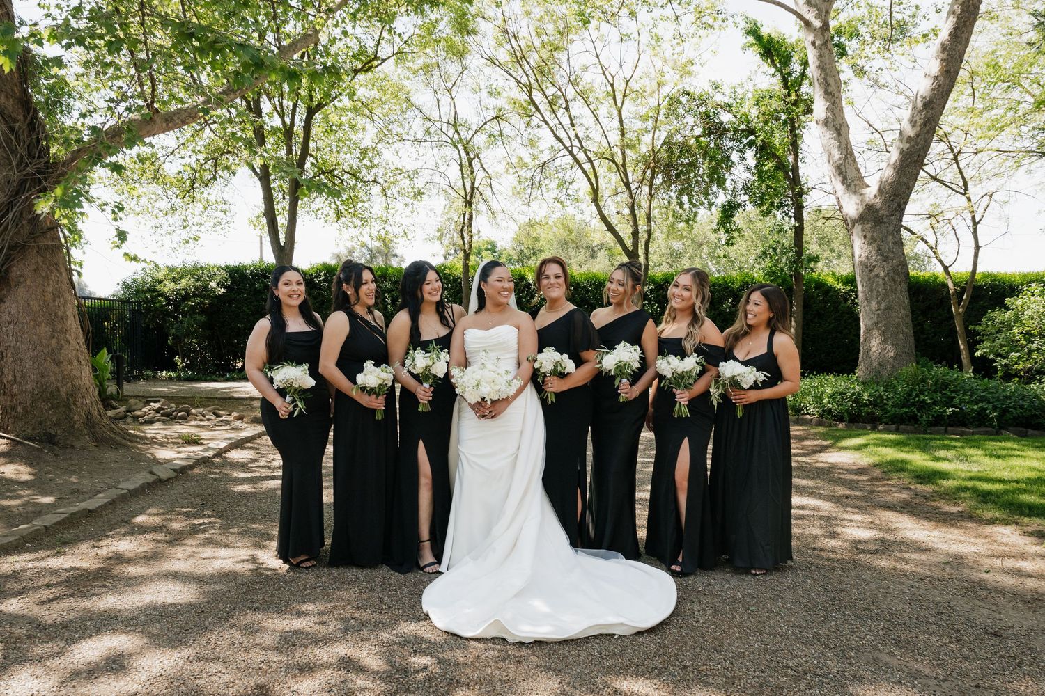 Bride in white gown poses with bridesmaids in black dresses holding white bouquets outdoors among lush green trees.