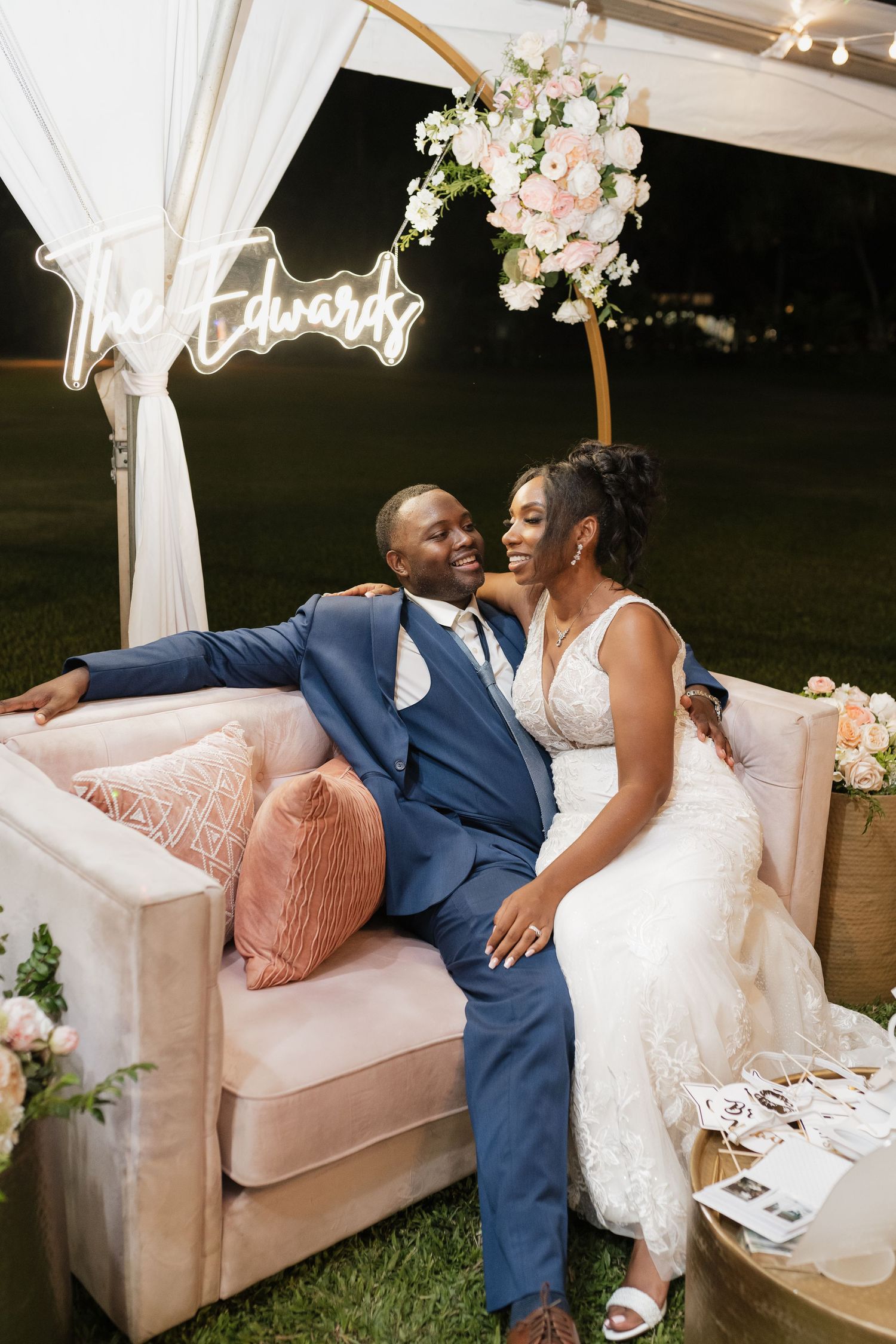 Couple relaxes on a pink velvet couch at an outdoor wedding reception with floral decorations.
