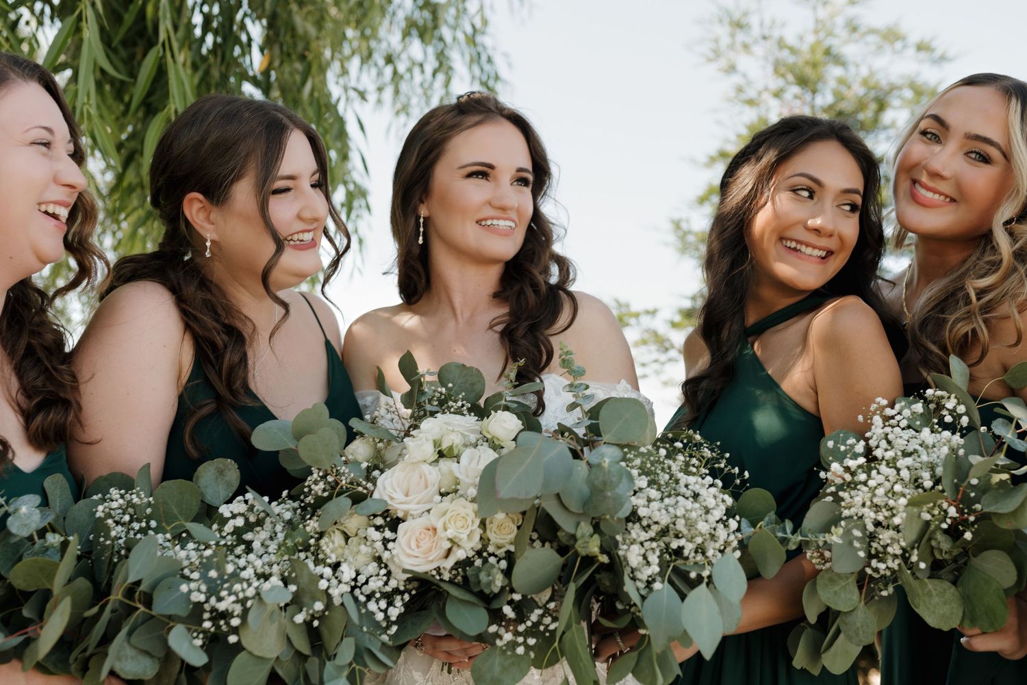 Wedding party holding bouquets of white flowers and eucalyptus against natural backdrop.