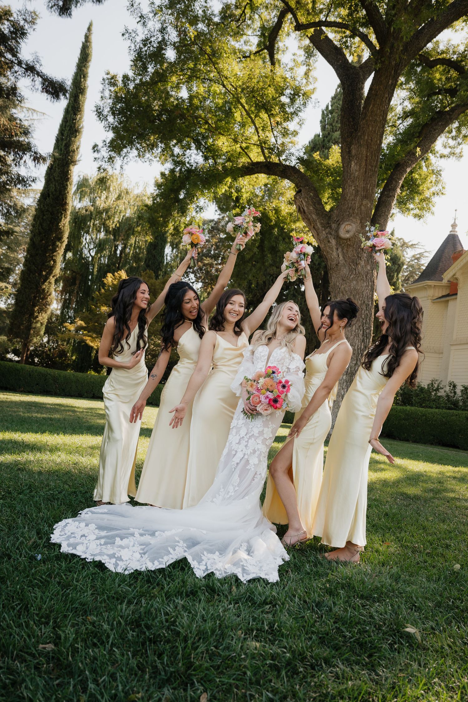 A group in cream-colored dresses and a white wedding gown pose joyfully together on a sunny lawn with trees in the background.