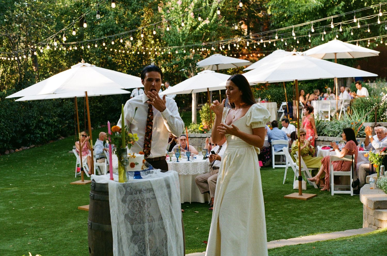 People celebrate at an outdoor garden wedding reception with white umbrellas, string lights and folding chairs on a green lawn.