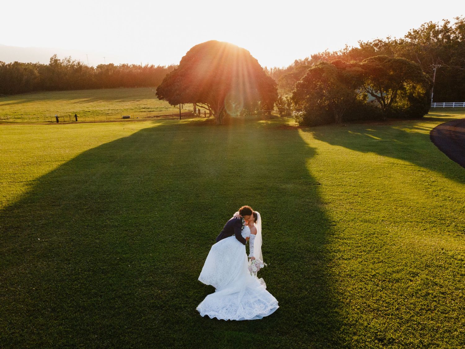 Bride and groom sharing a romantic dip kiss on a lush green lawn at golden sunset hour.