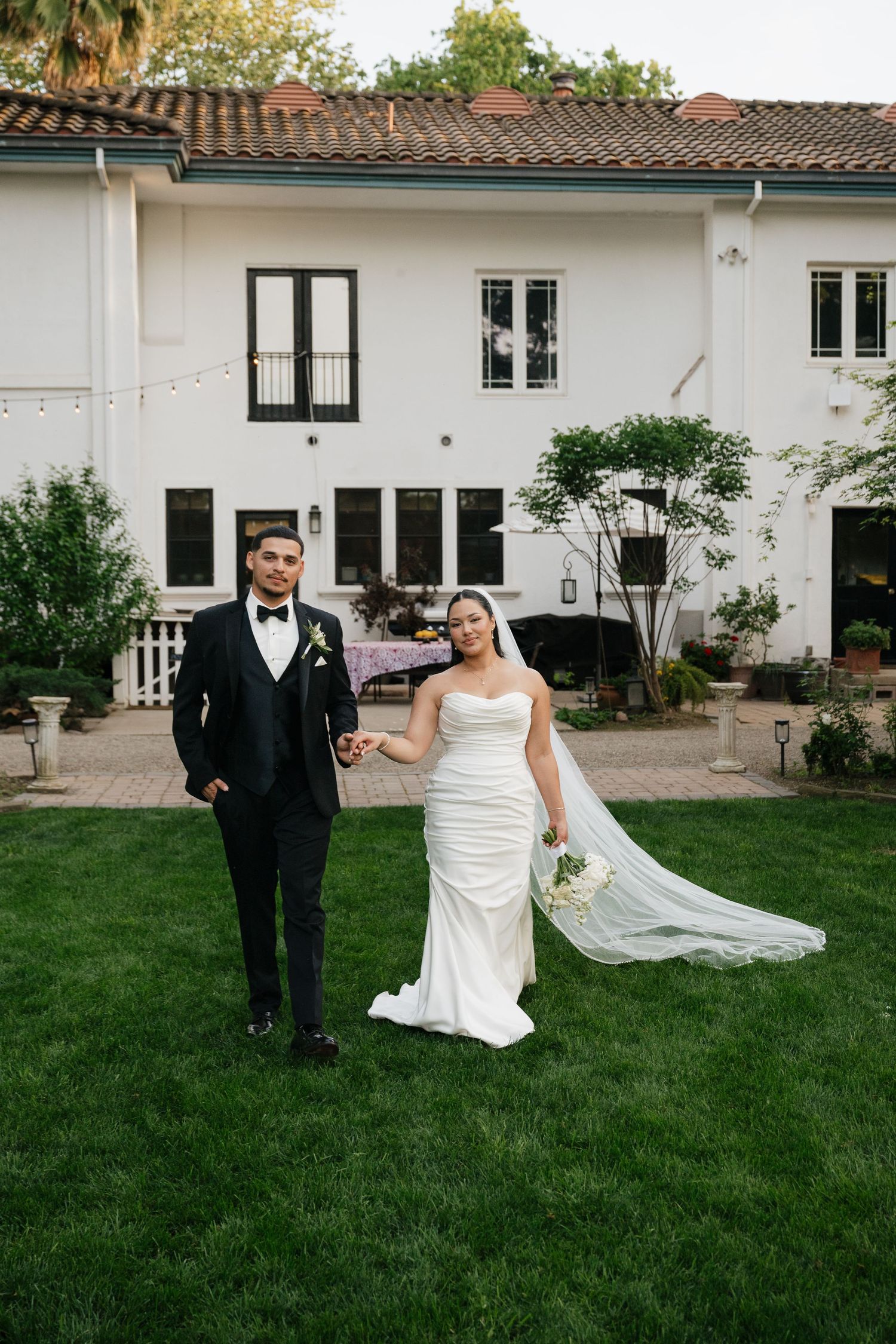 Bride and groom holding hands on green lawn in front of elegant white Spanish-style villa at outdoor wedding.