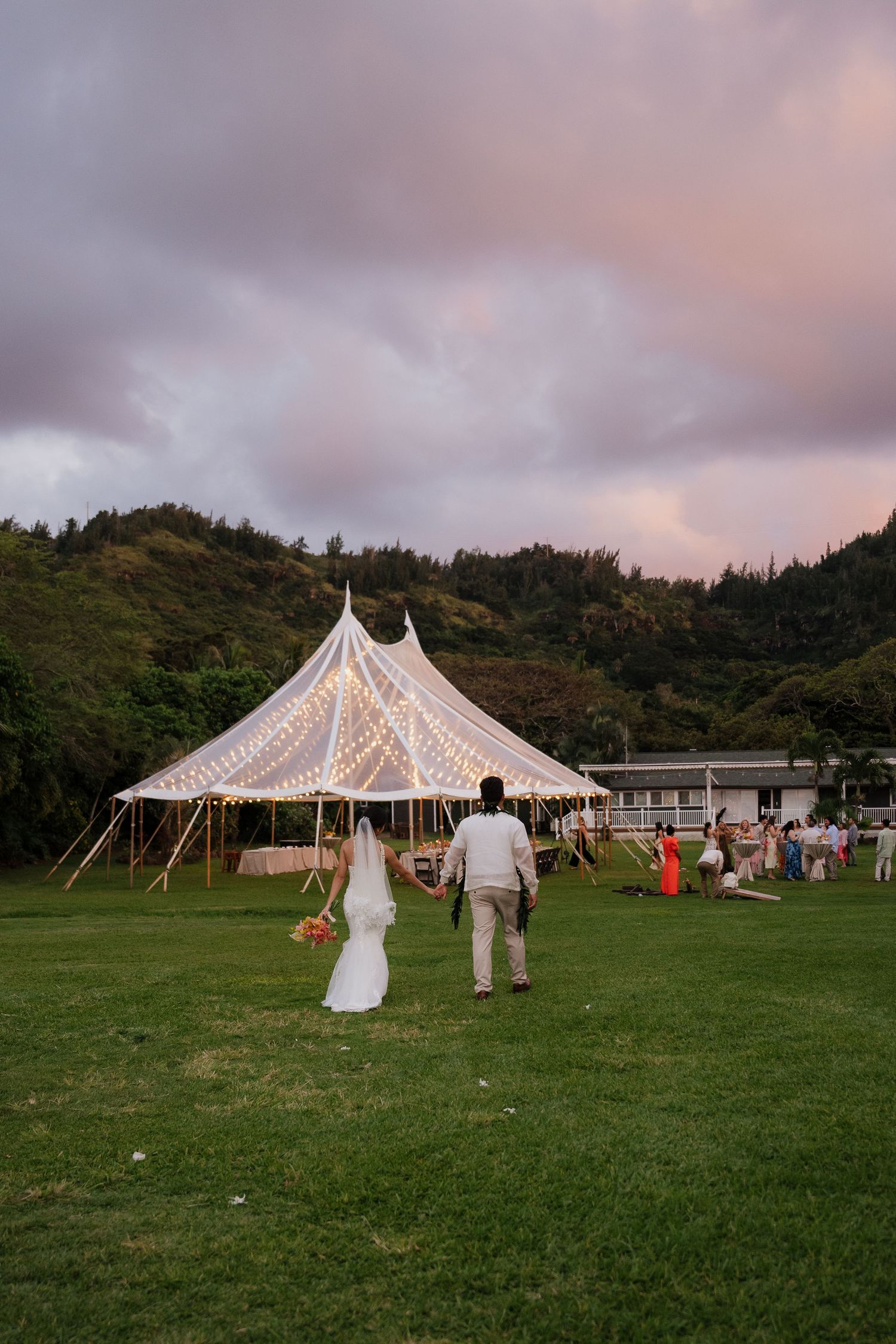 Bride and groom walk toward a glowing tent at sunset with lush green hills in the background at an outdoor wedding.