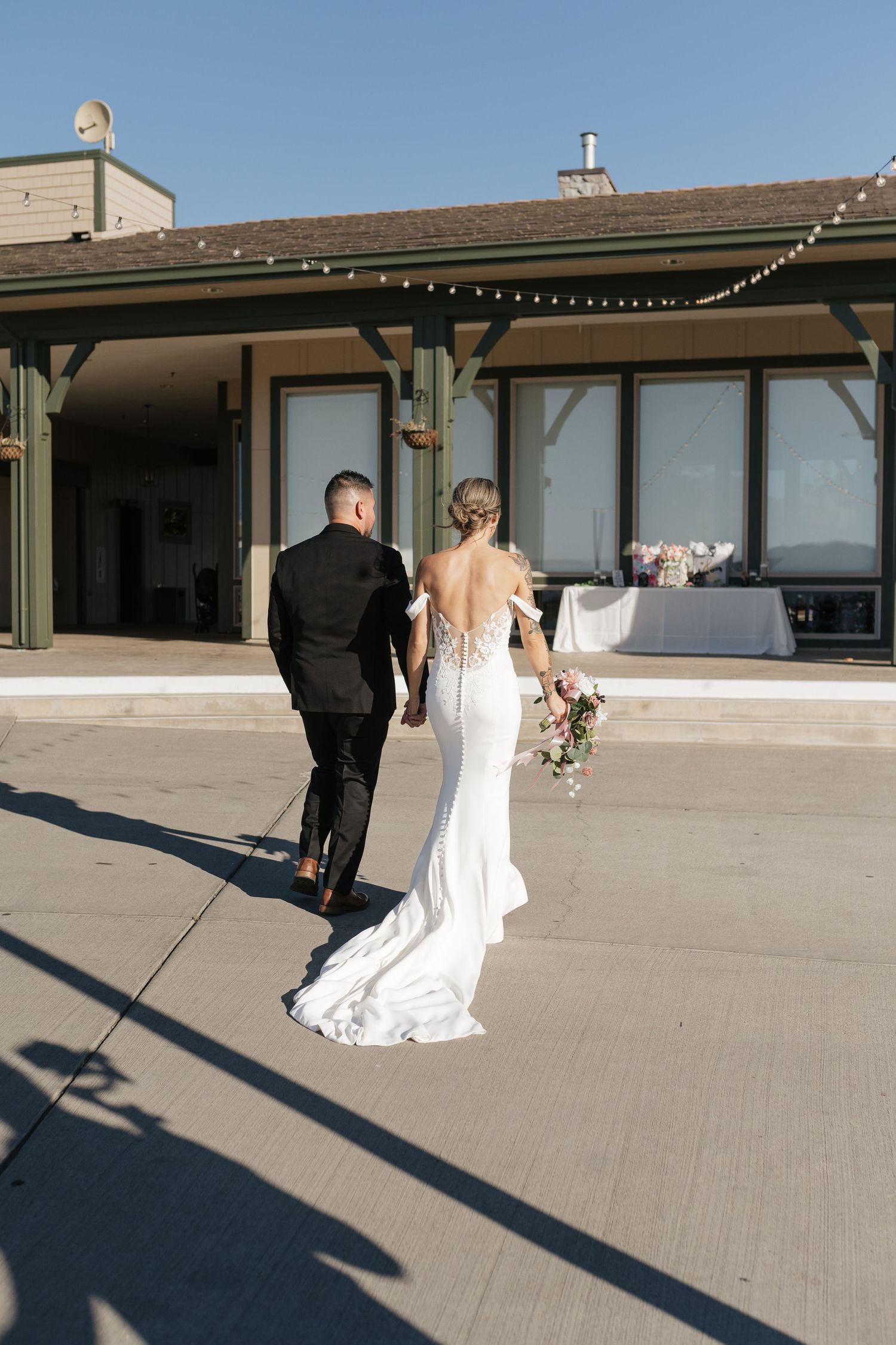 Wedding couple walks along concrete path outside modern building with string lights.