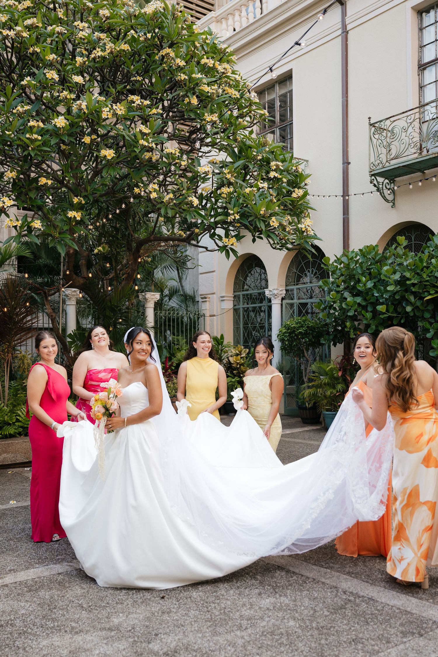 Bride in white gown holds bouquet while bridesmaids in colorful dresses lift her train outdoors near elegant building.