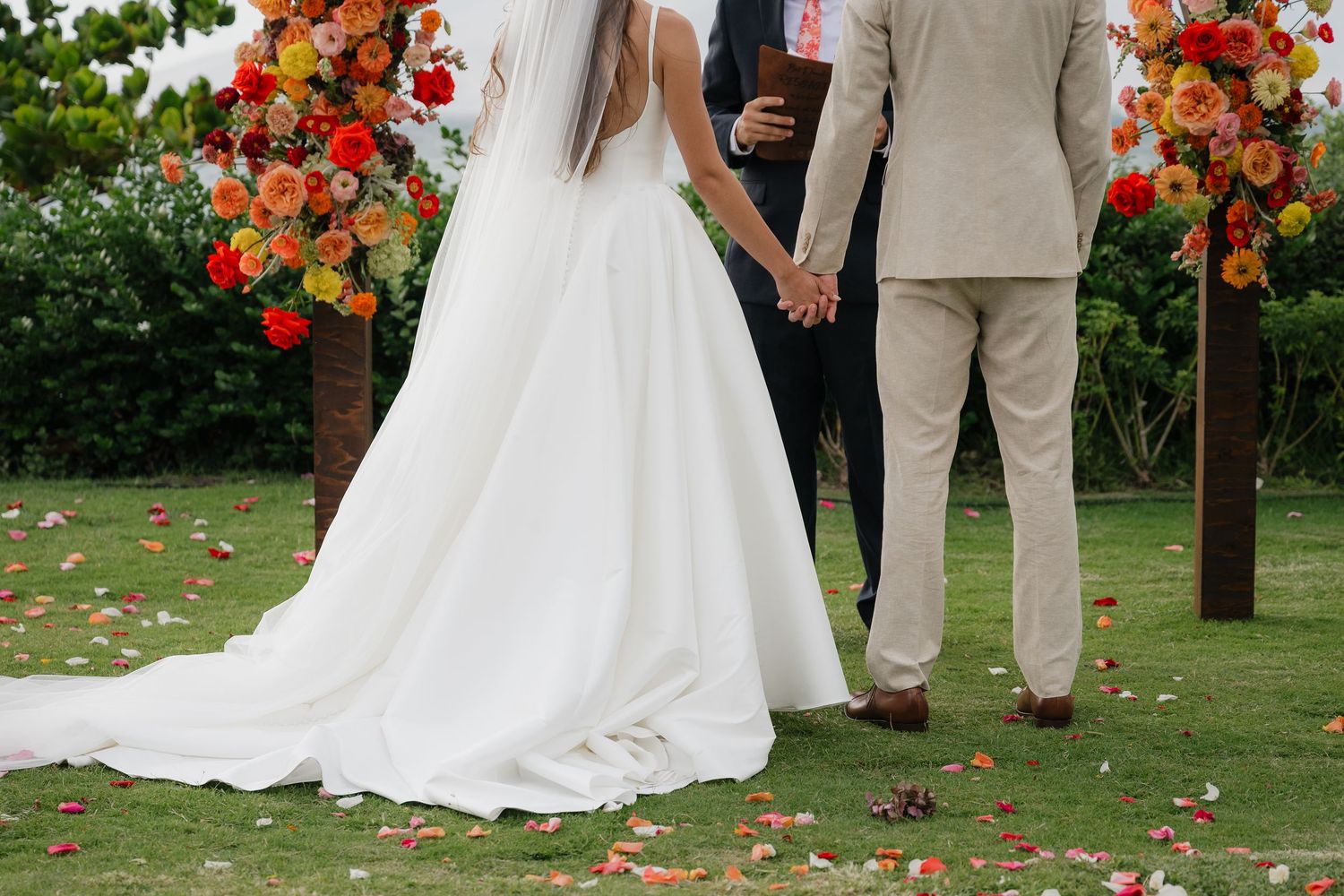 A couple holds hands at an outdoor garden wedding ceremony with vibrant orange floral arrangements and scattered petals.