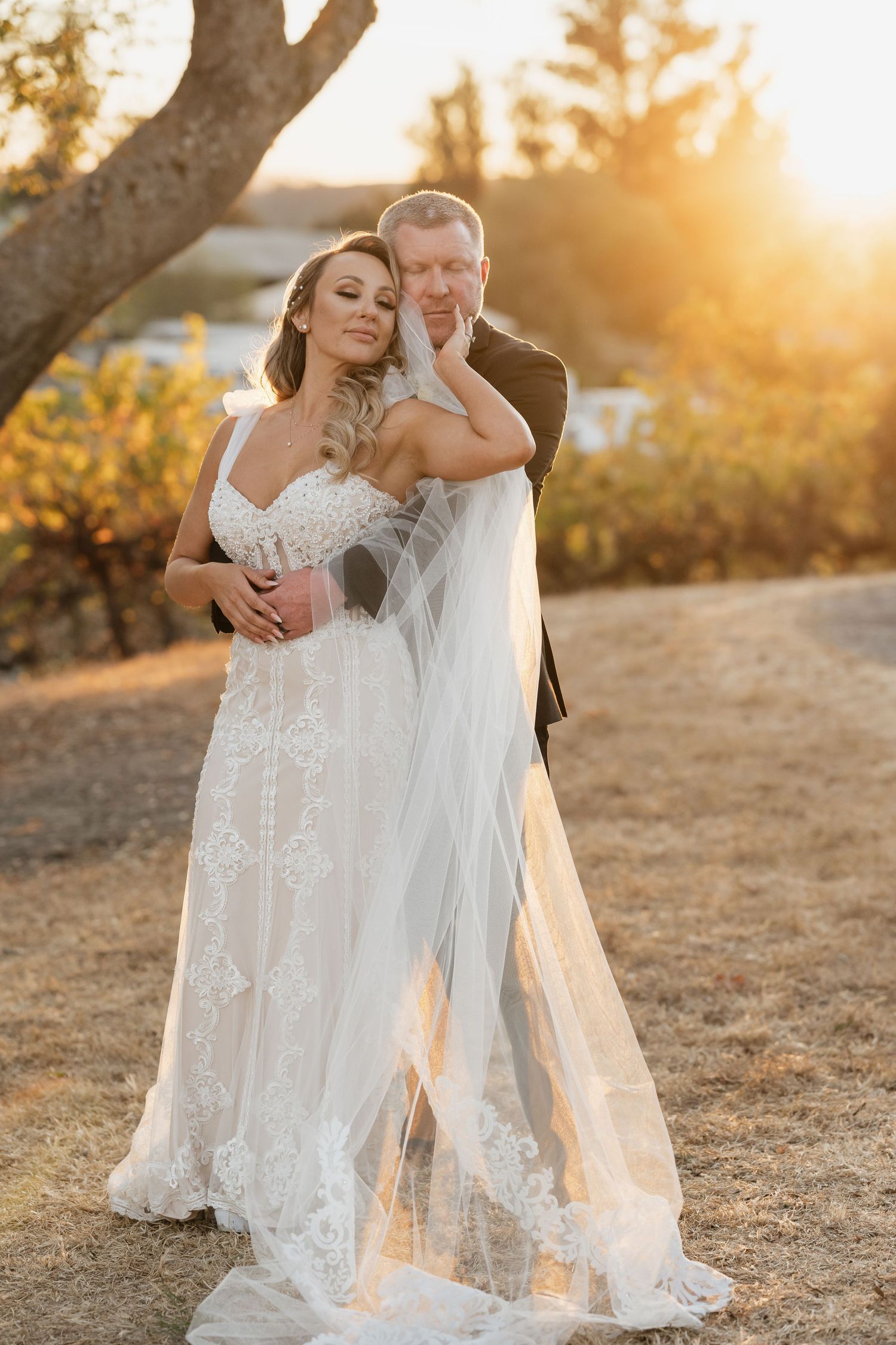 A romantic wedding photo of a couple embracing in golden sunset light with a flowing lace dress and veil.