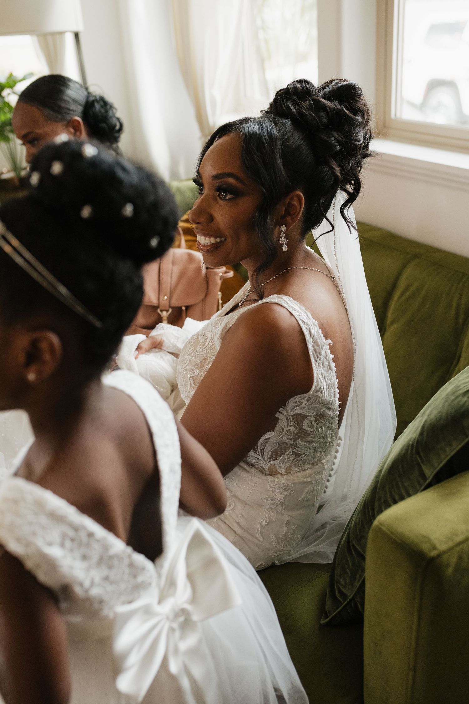 Bride in a stunning white lace wedding dress with bow detail sits on a green couch.