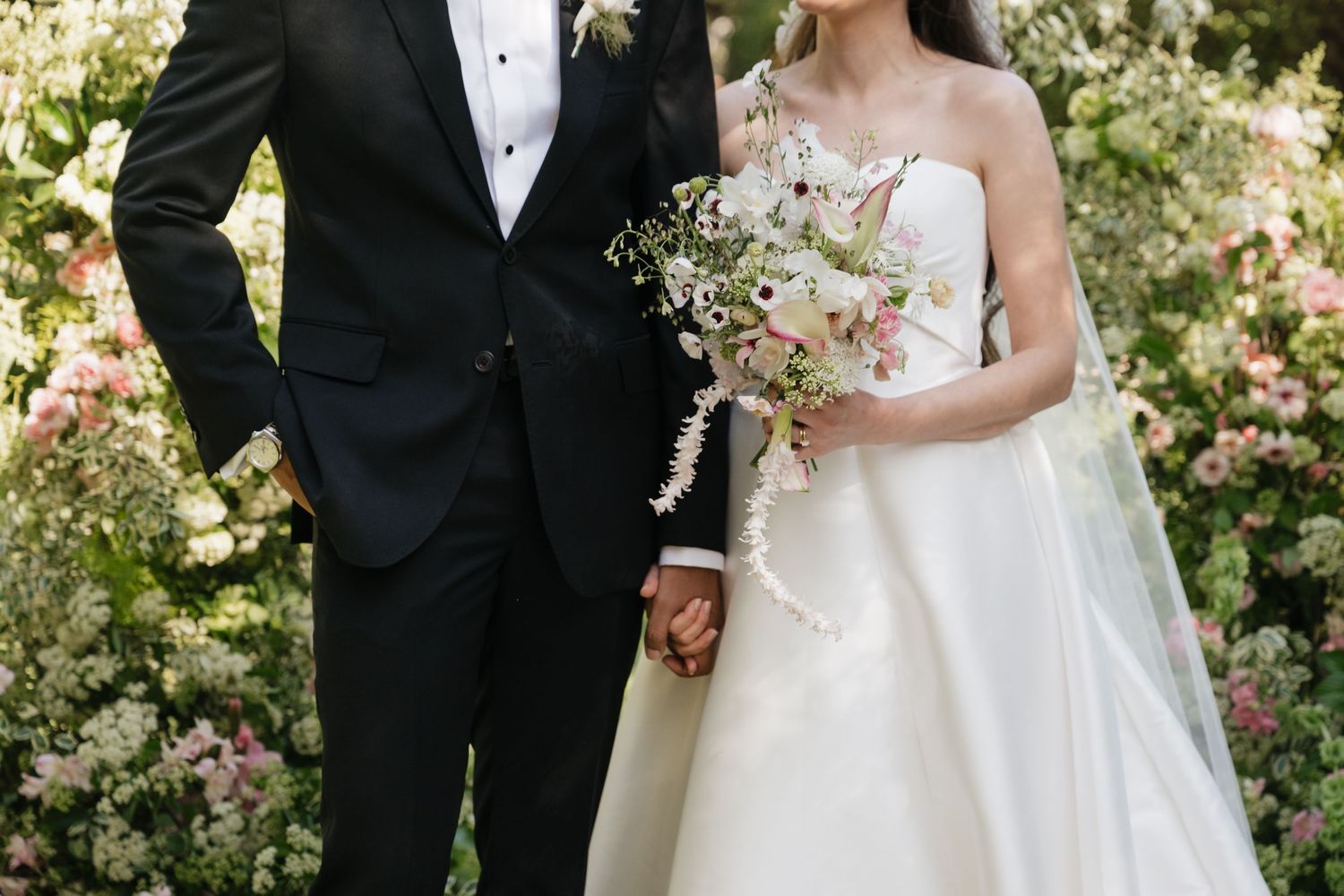 A bride and groom hold hands in a lush garden, she in a white gown with bouquet, he in a black tuxedo.