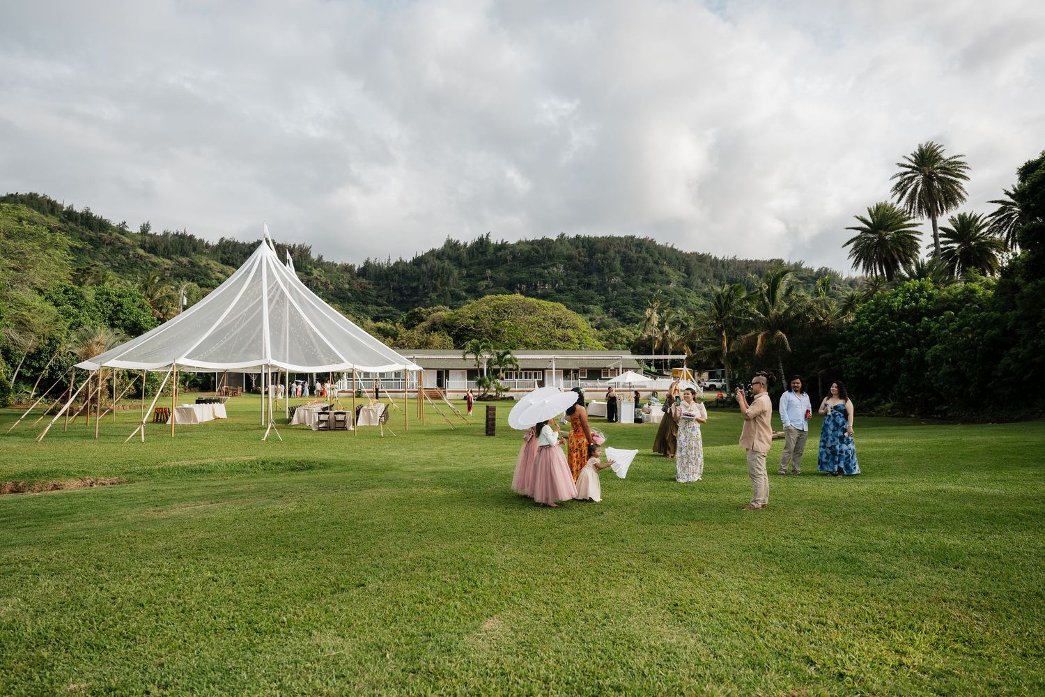 Wedding guests gather on lush green lawn near elegant white tent venue surrounded by tropical hills and palm trees.