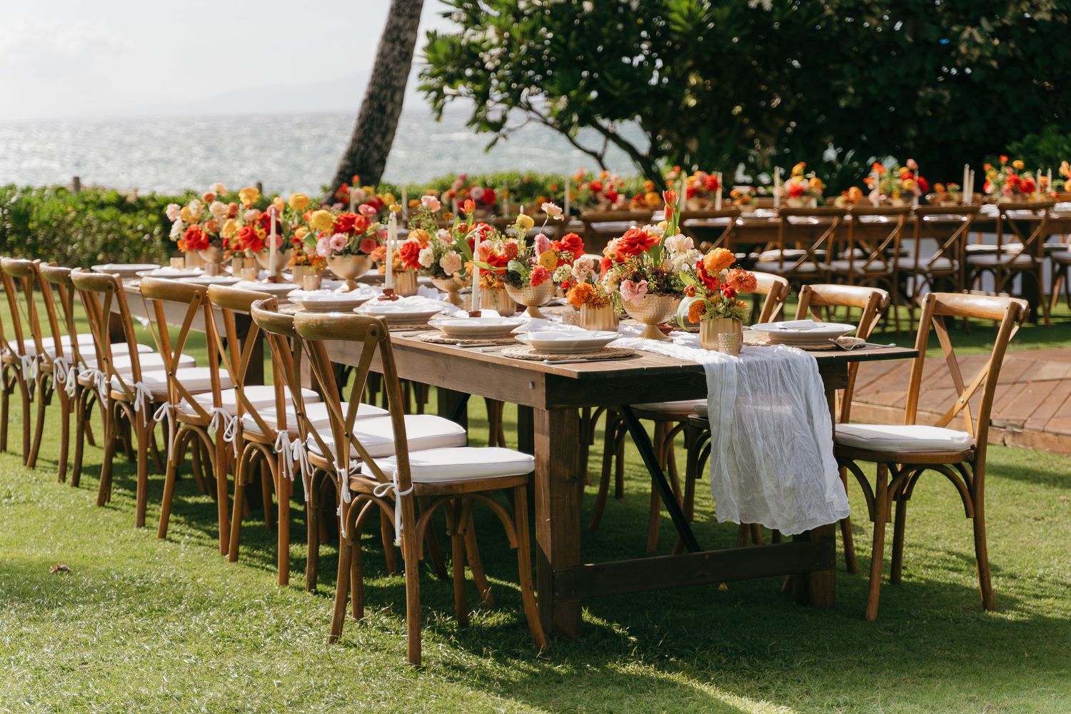 Rustic outdoor wedding reception setup with wooden cross-back chairs and white tablecloths overlooking a scenic landscape.