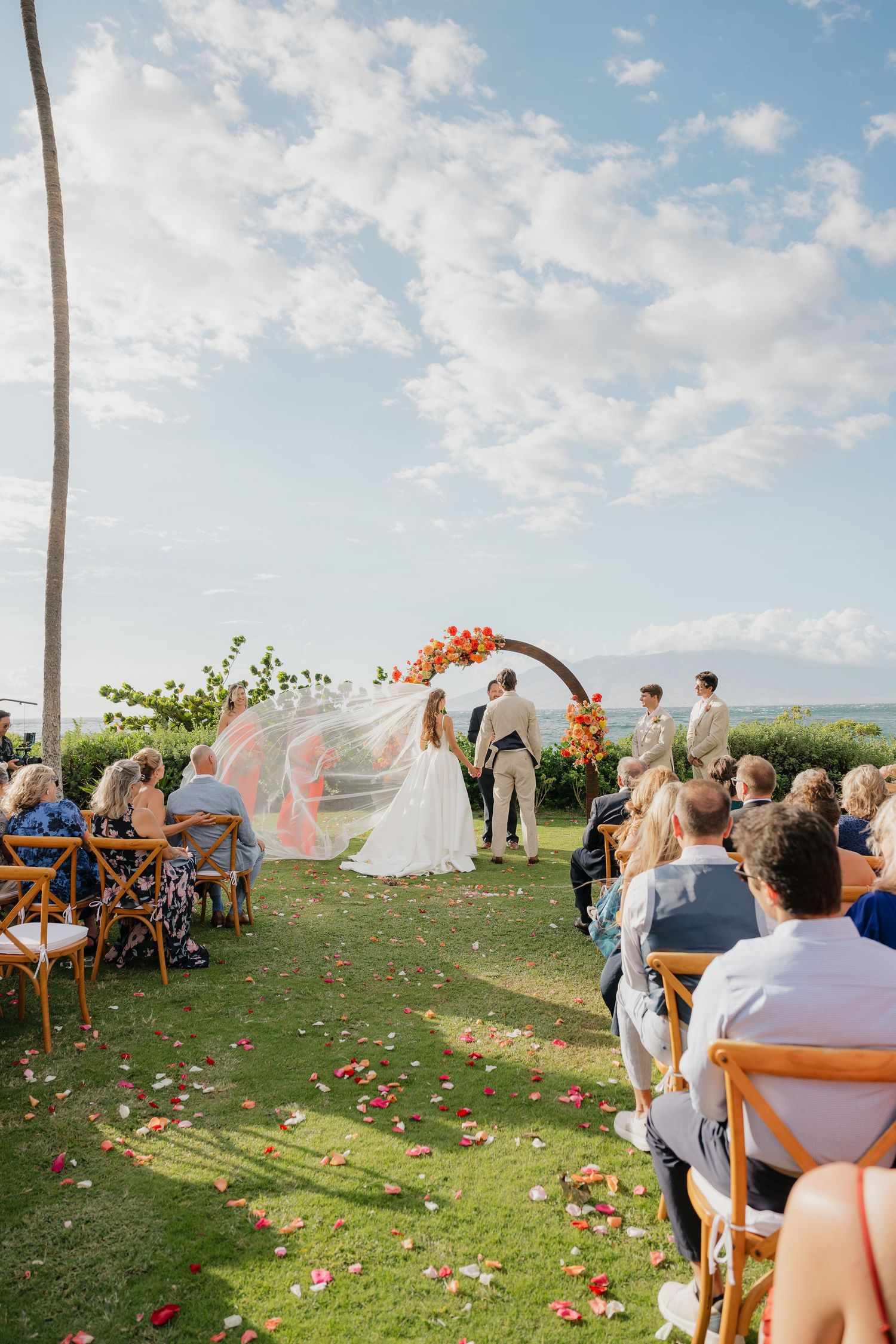 Beachside wedding ceremony setup with wooden benches, orange floral arrangements, and palm trees in the background.