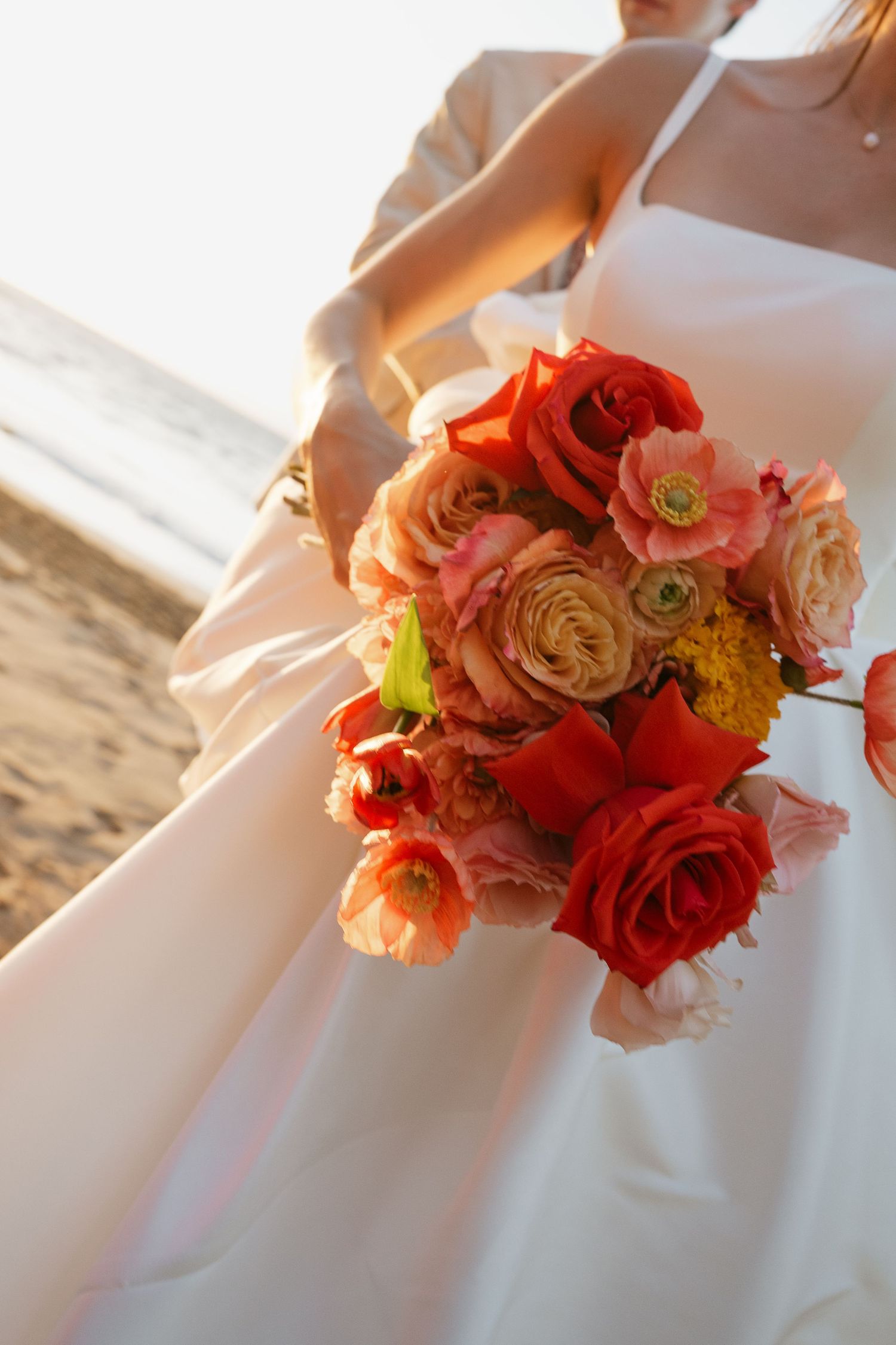 A coral and peach colored bridal bouquet with ranunculus flowers held against a white wedding dress on a beach.