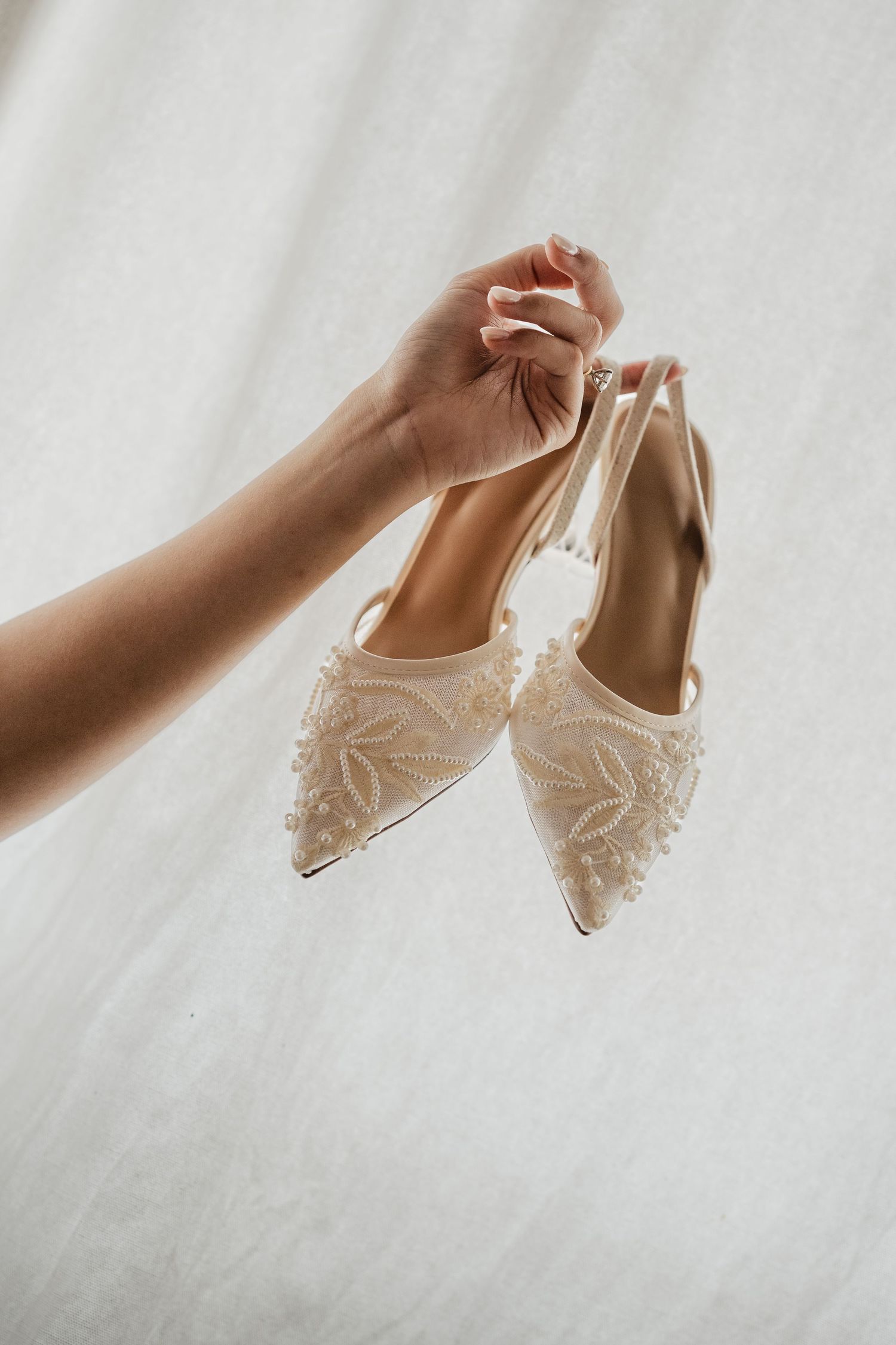 A hand holds elegant ivory bridal heels adorned with delicate pearl and lace floral embroidery against a white background.