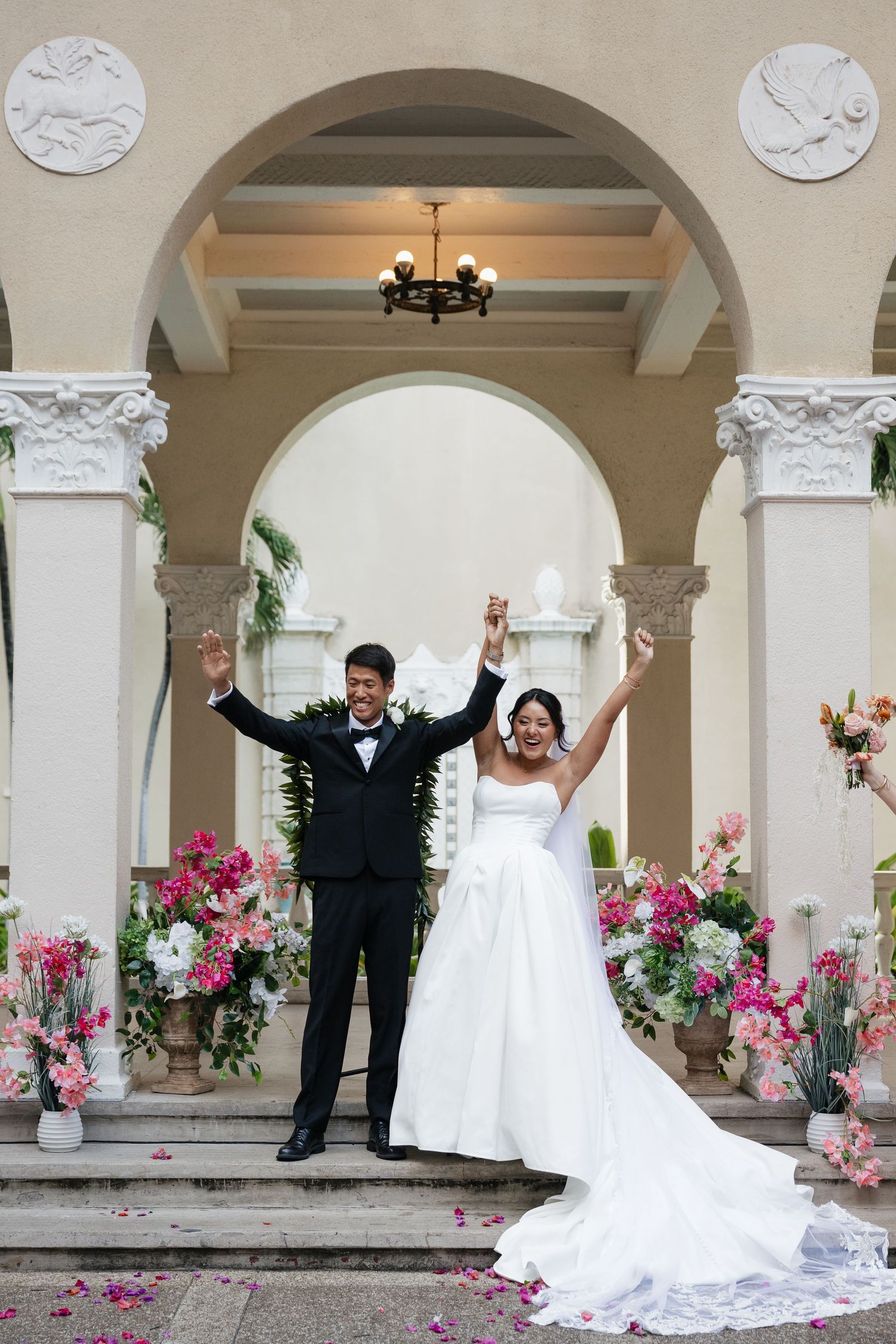 Joyful bride and groom raising hands at elegant wedding ceremony venue with pink floral arrangements and arched columns.