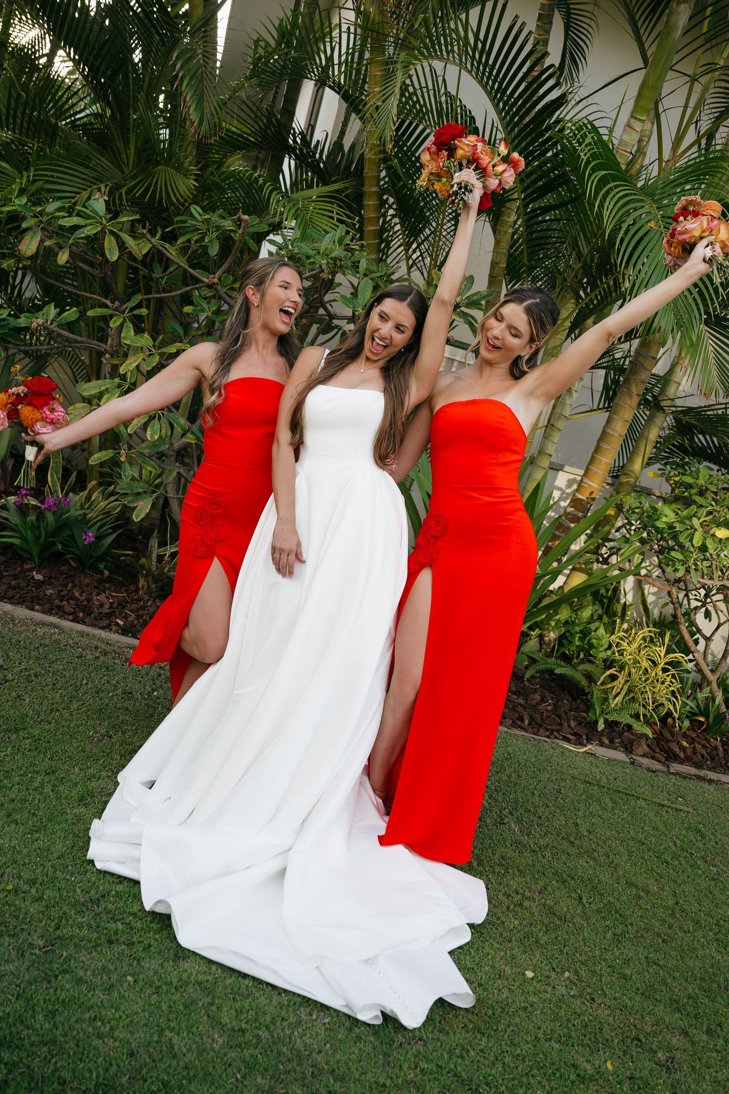 Three women in red and white dresses pose playfully on a lawn with tropical plants and palm trees in the background.
