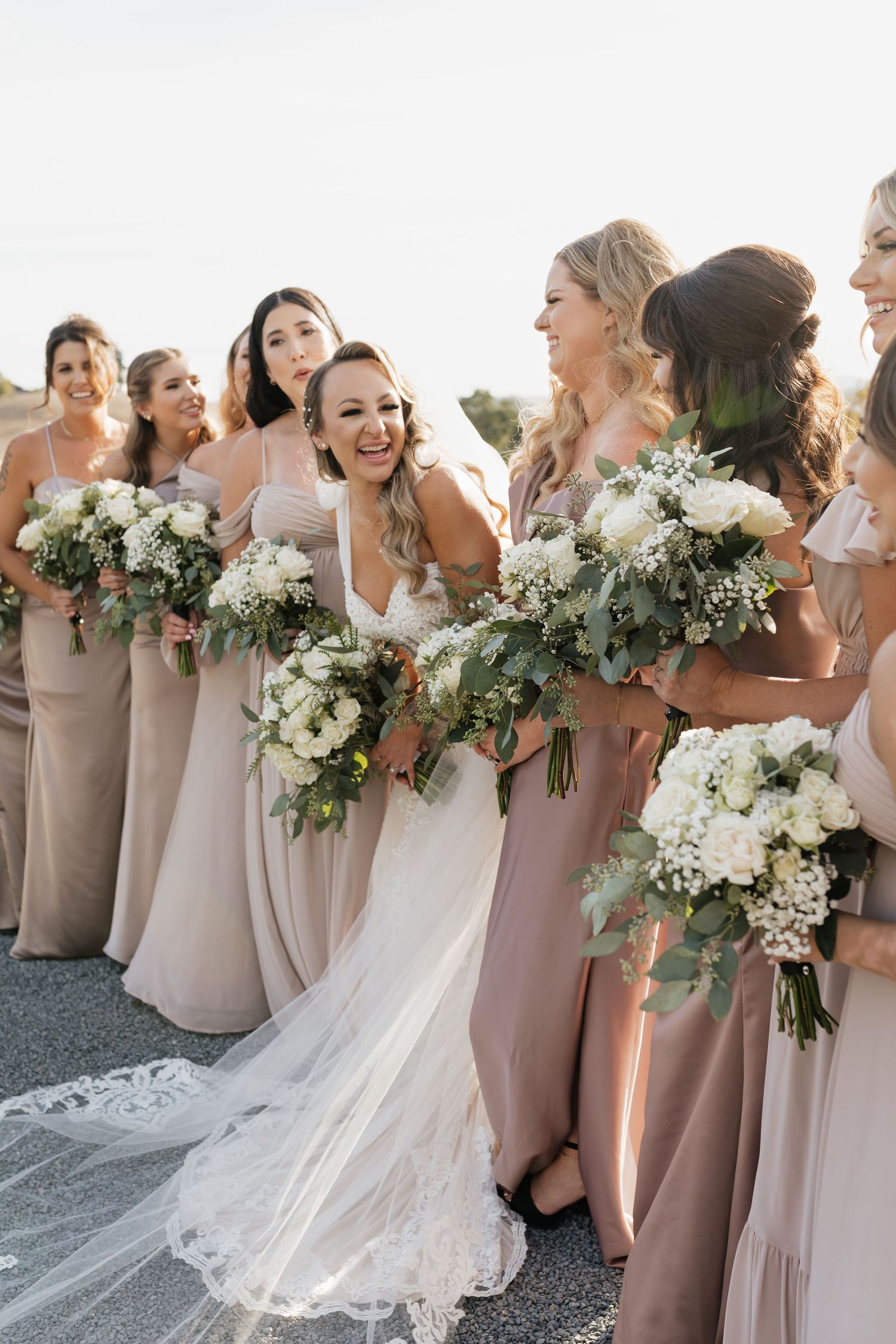 Bridesmaids in blush dresses hold white and green bouquets while laughing together.