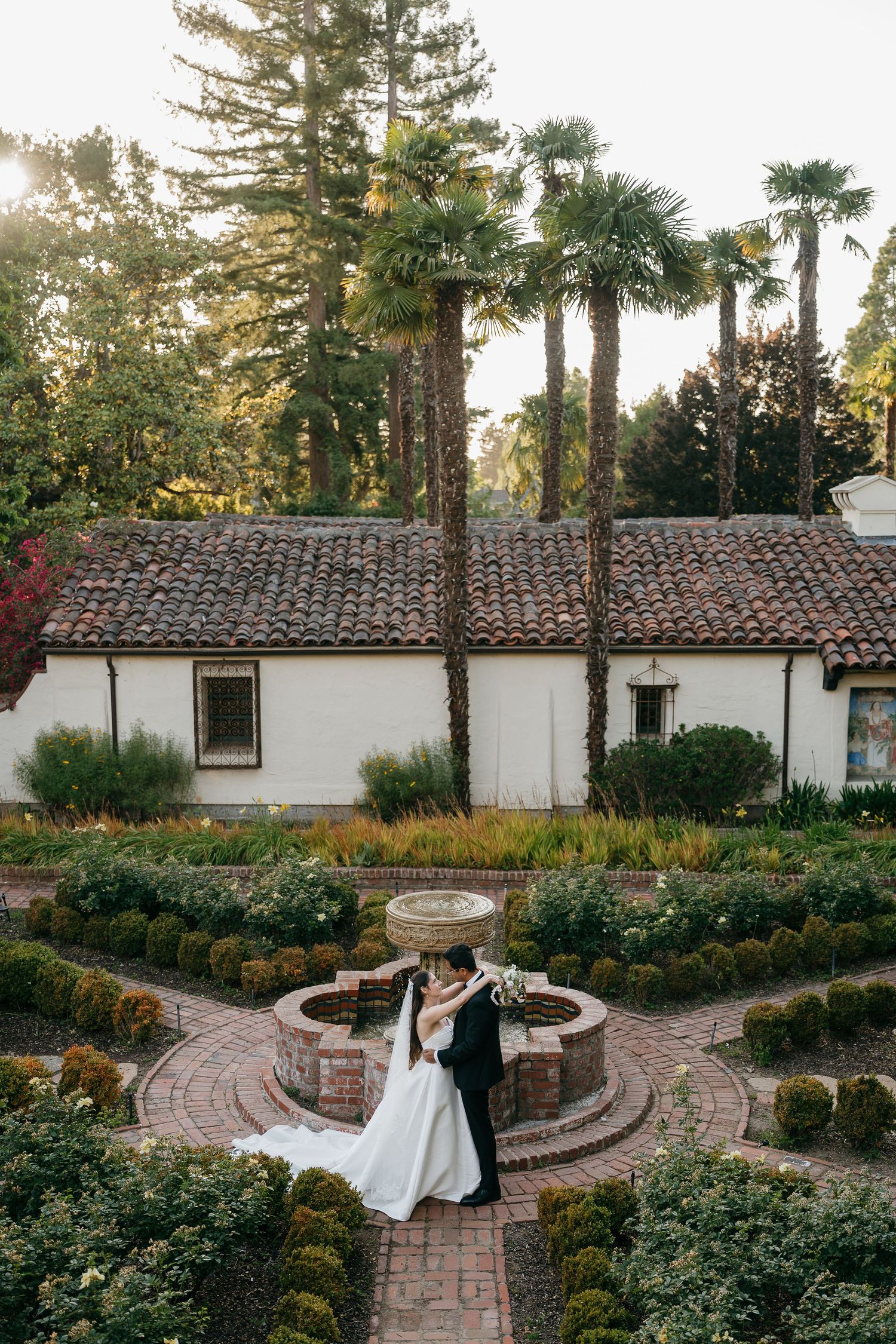 Bride and groom embrace in a formal garden with a stone fountain, Spanish-style building, and tall palm trees at golden hour.