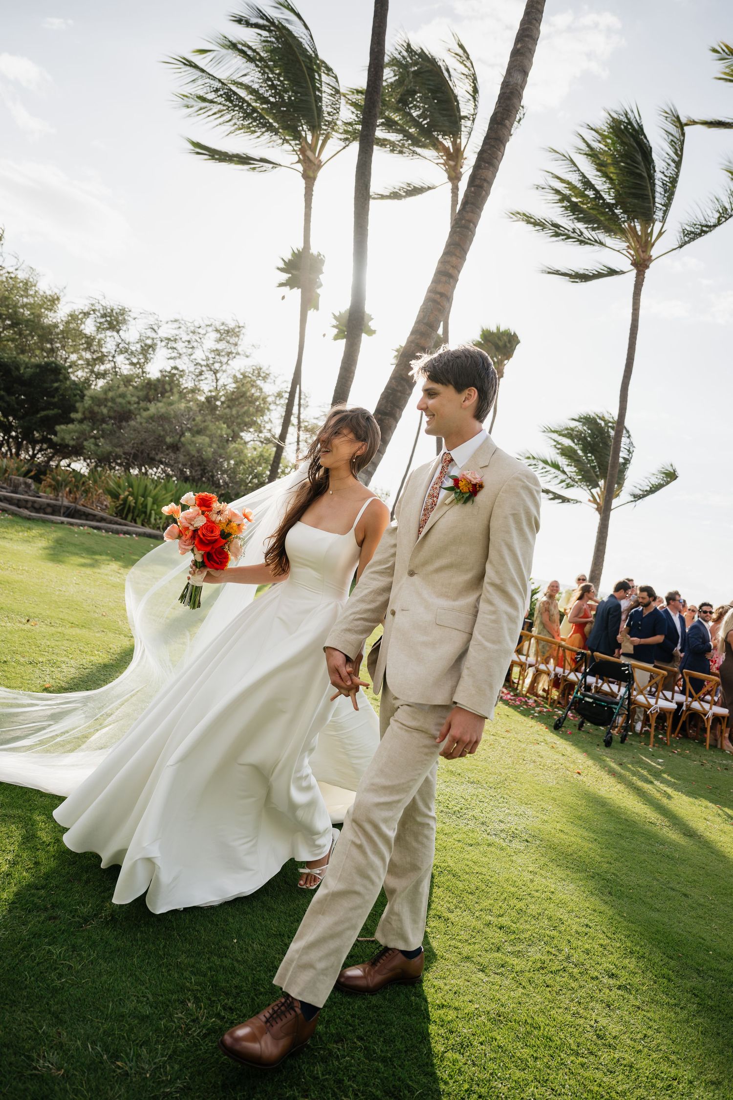 A couple celebrates their wedding on a breezy oceanfront lawn with palm trees swaying in the background.