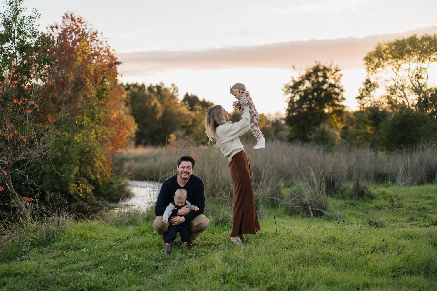 A couple stands in a golden meadow at sunset with trees silhouetted against the warm sky.