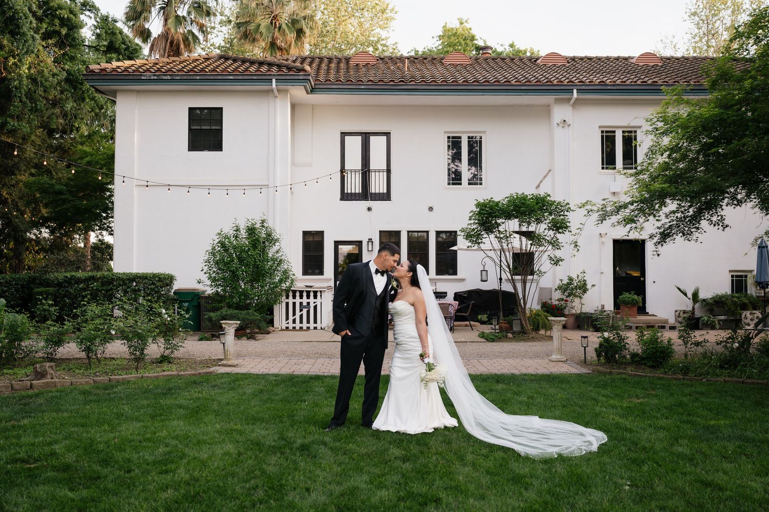 Bride and groom share a romantic kiss on lush green lawn in front of elegant white wedding venue.
