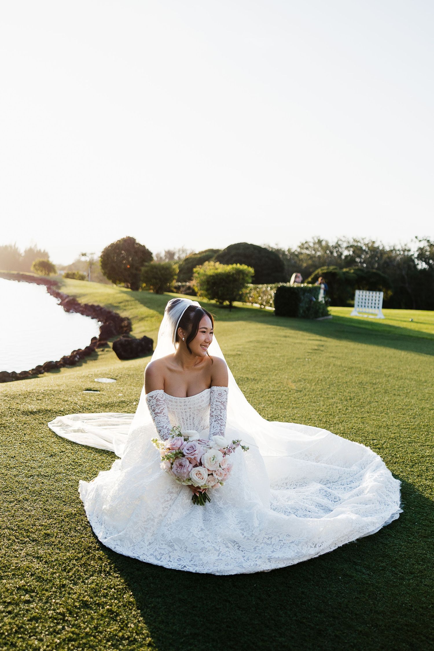 Bride in off-shoulder lace gown sits on golf course lawn holding pink bouquet at golden hour.